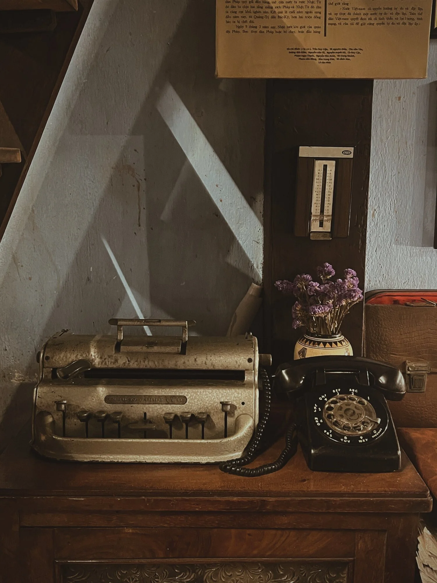 Vintage typewriter on a wooden surface with a rotary telephone, a vase with purple flowers, and a crocheted bag.