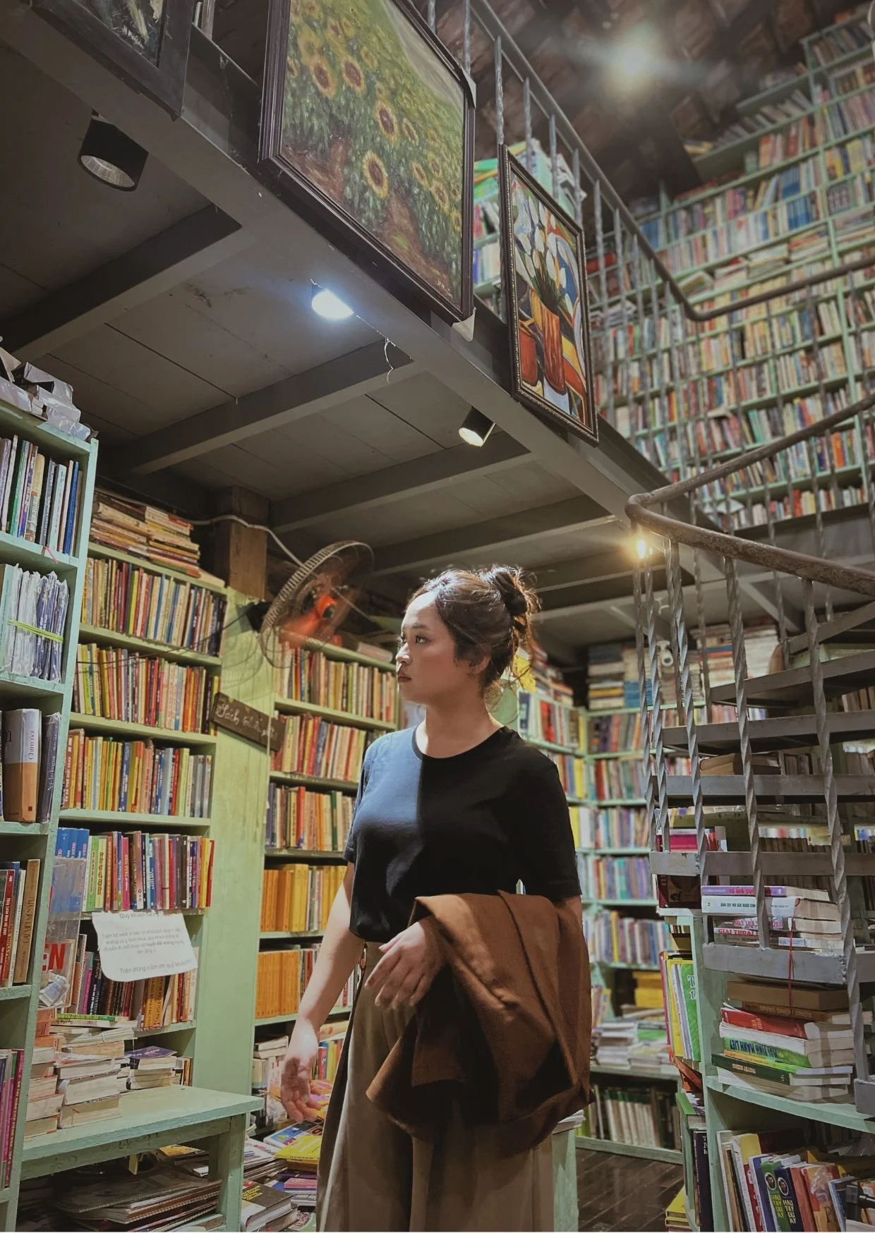 A woman shopping for books in a bookstore. She is holding a brown jacket and wearing a black t-shirt, with a room full of bookshelves behind her.