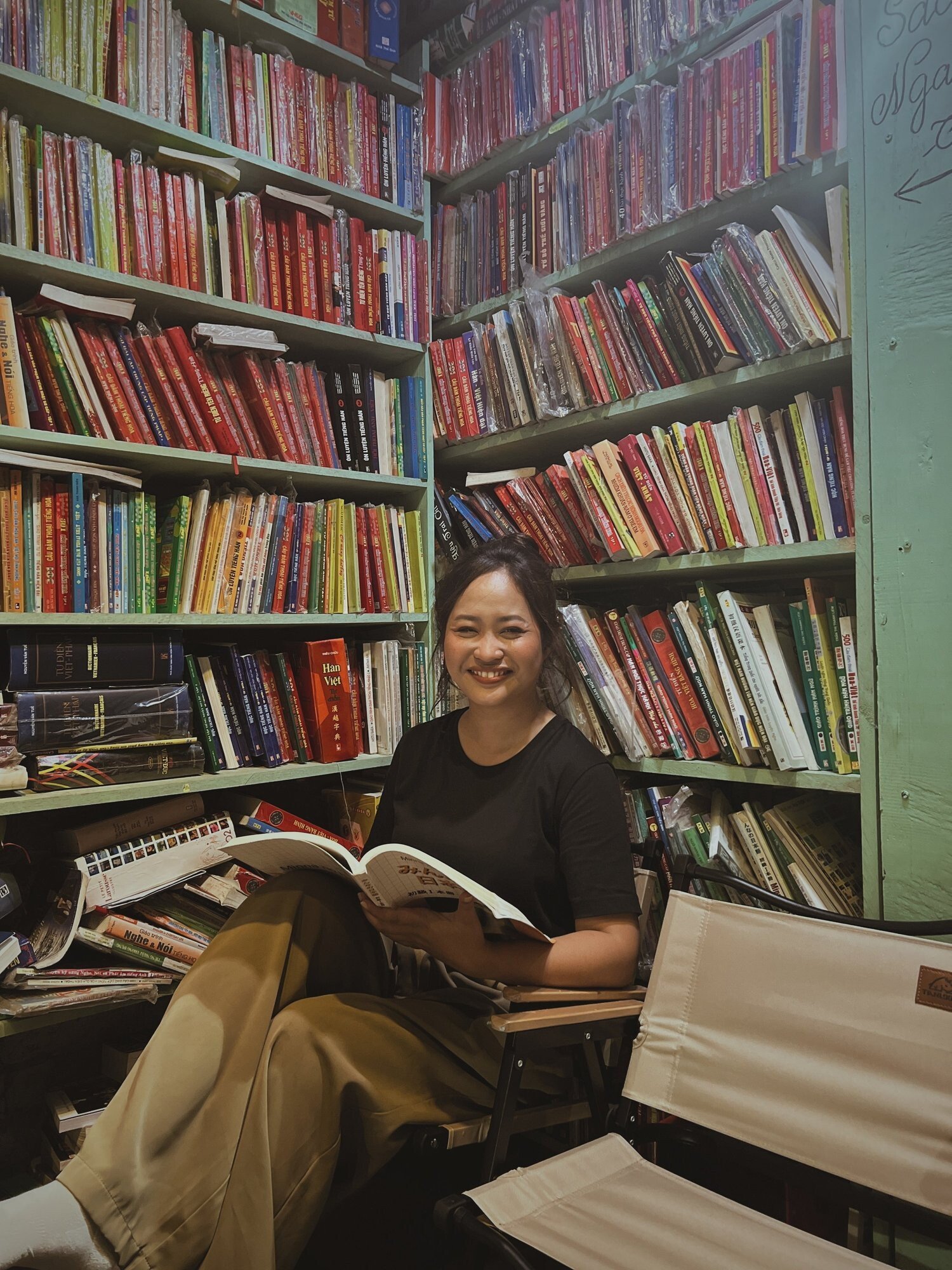 A smiling woman with dark hair sitting in a bookstore or library, holding an open book, surrounded by tall bookshelves filled with colorful books.