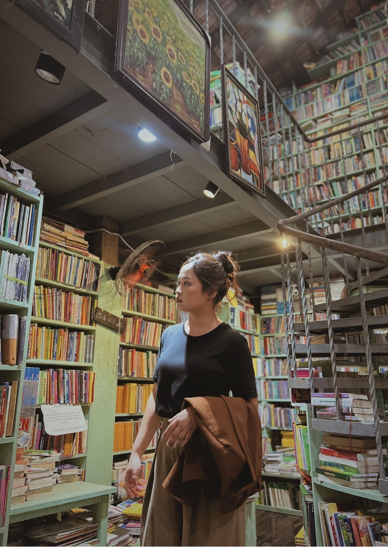 A woman in a black t-shirt shopping for books at a bookstore or library with tall shelves filled with books. She is holding a brown coat or jacket and looking at the books. There are paintings hanging from the ceiling.