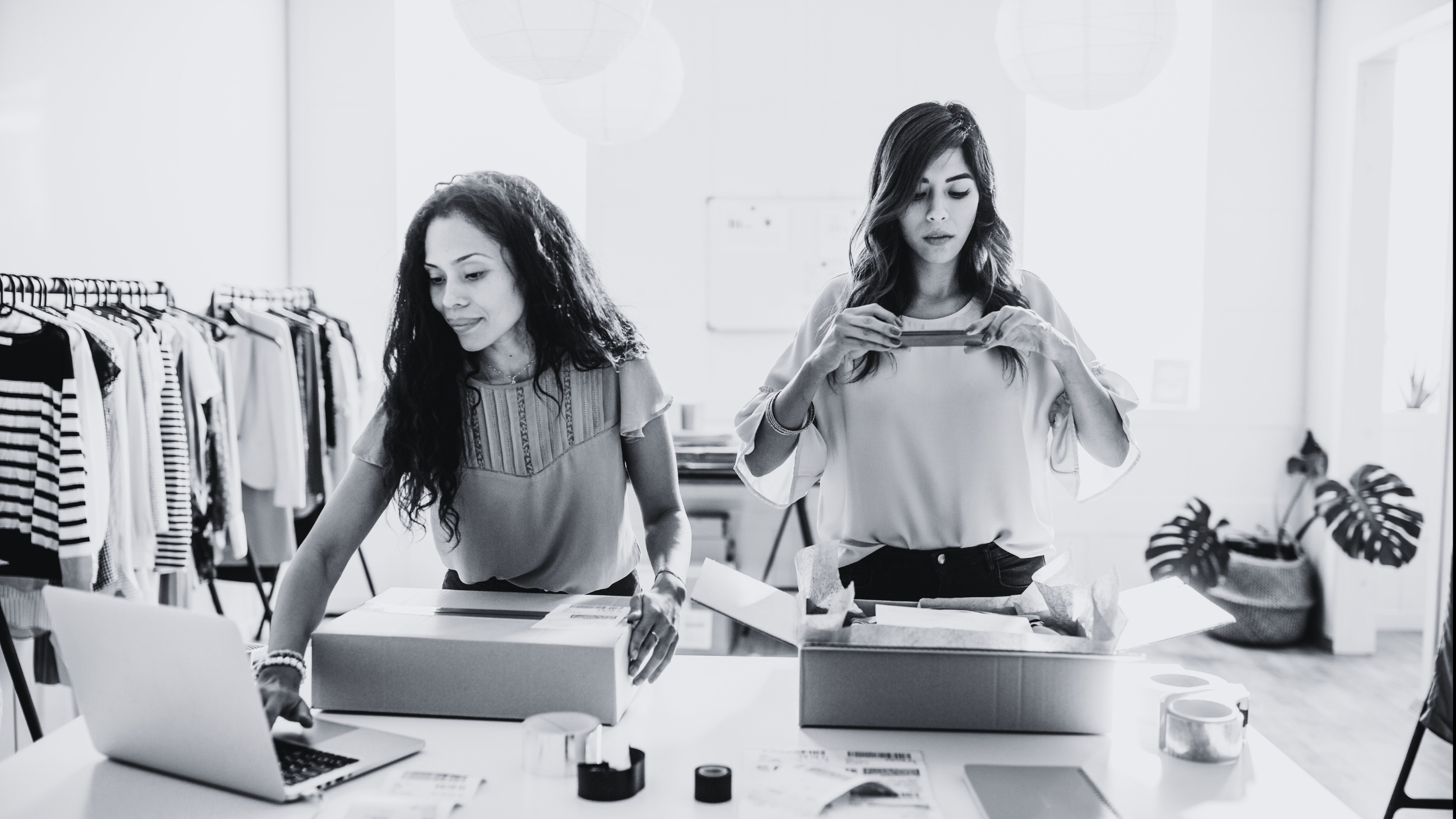 Two women in a modern, well-lit office or workspace, unpacking products or materials on a table, with clothing racks in the background.
