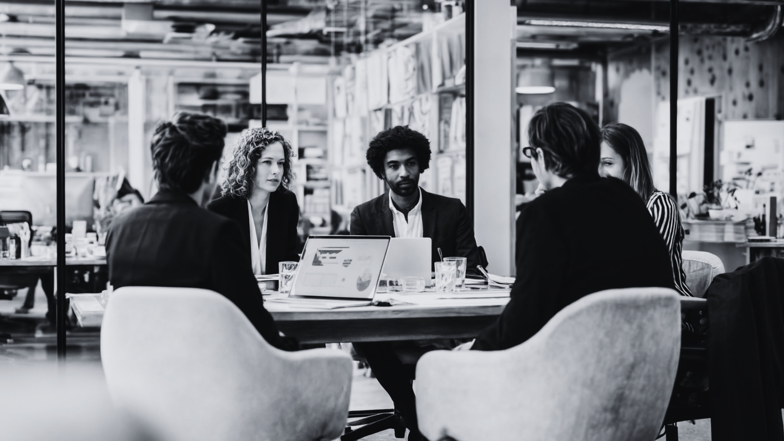 Group of five people having a meeting at a conference table in a modern office with bookshelf background, black and white photo.
