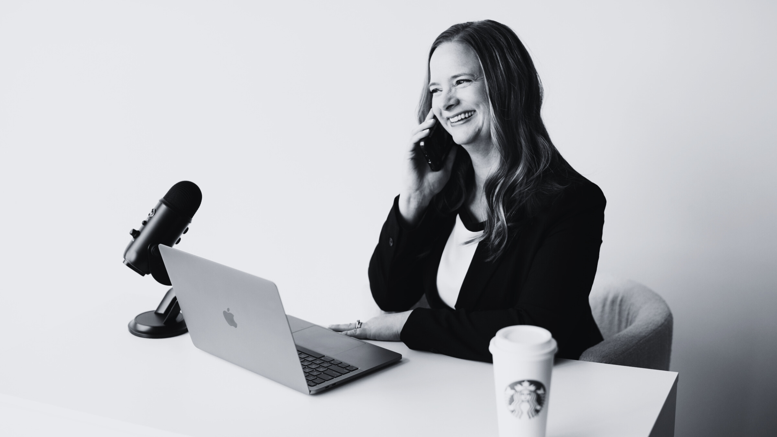 A woman smiling on a phone call, sitting at a desk with a laptop, microphone, and Starbucks cup.