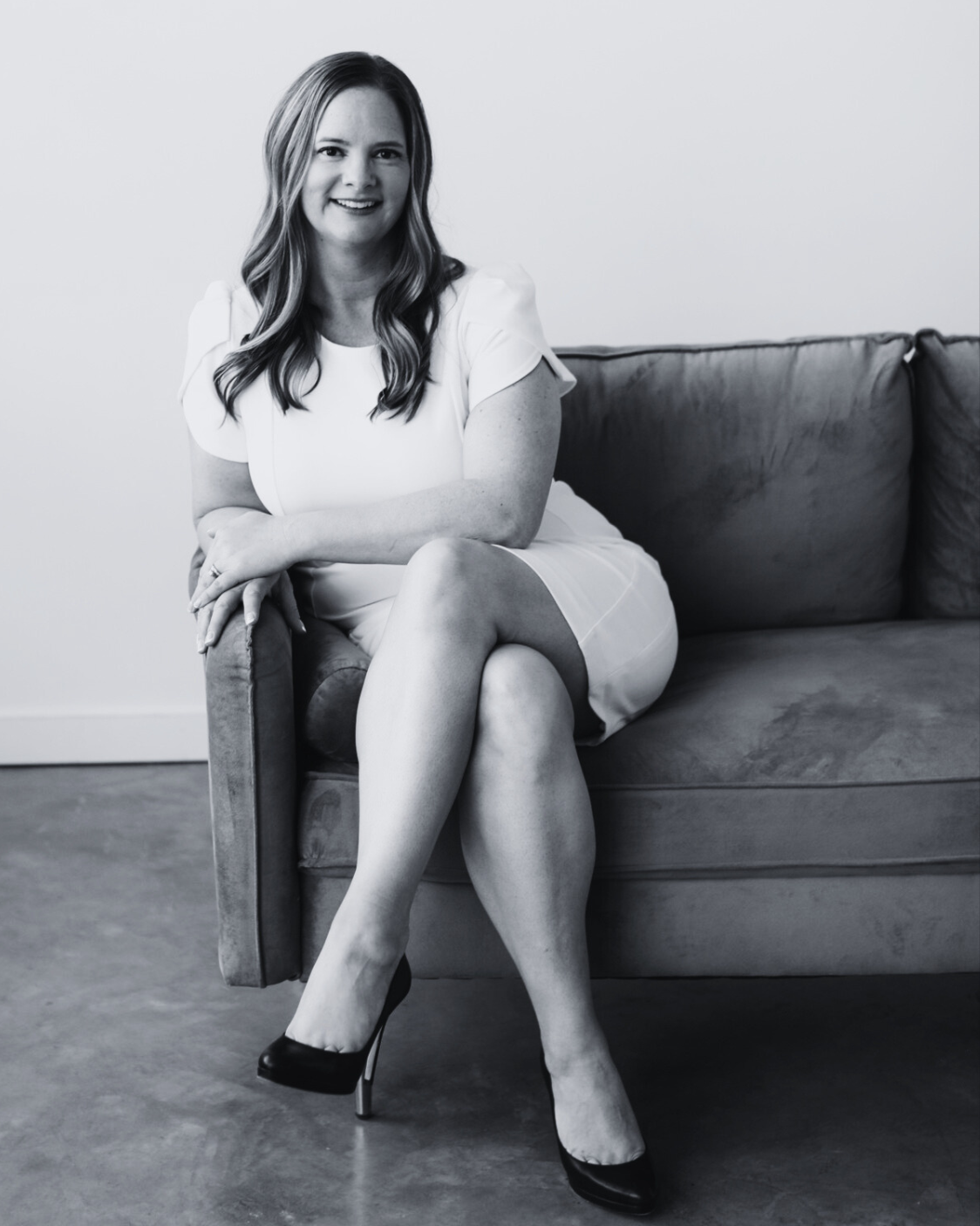 Jen Dunn Calgary Business Advisor iwith long hair sitting on a sofa, wearing a white dress and high heels, smiling at the camera.