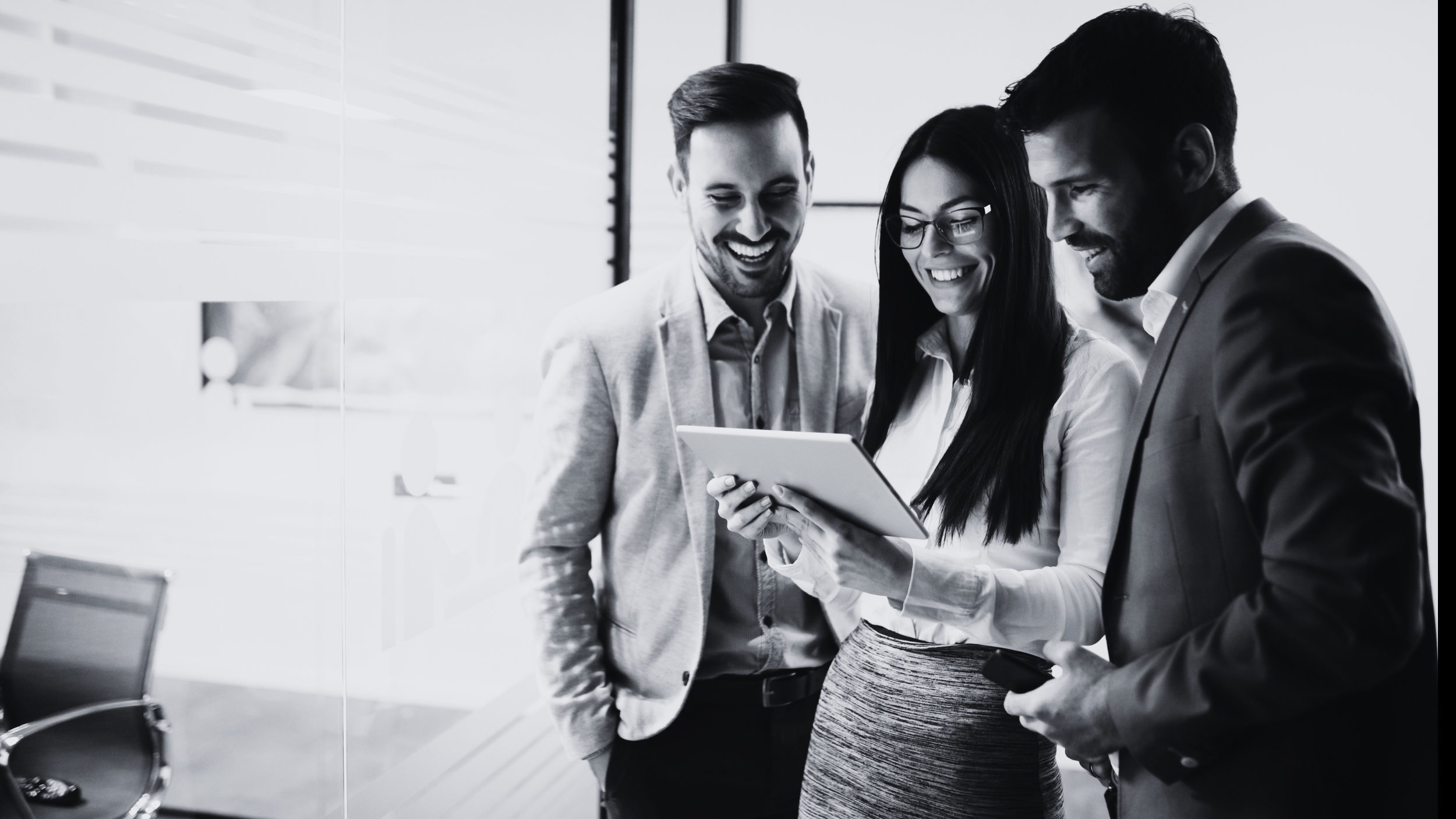 Three business professionals standing together in a modern office, looking at a tablet and smiling.