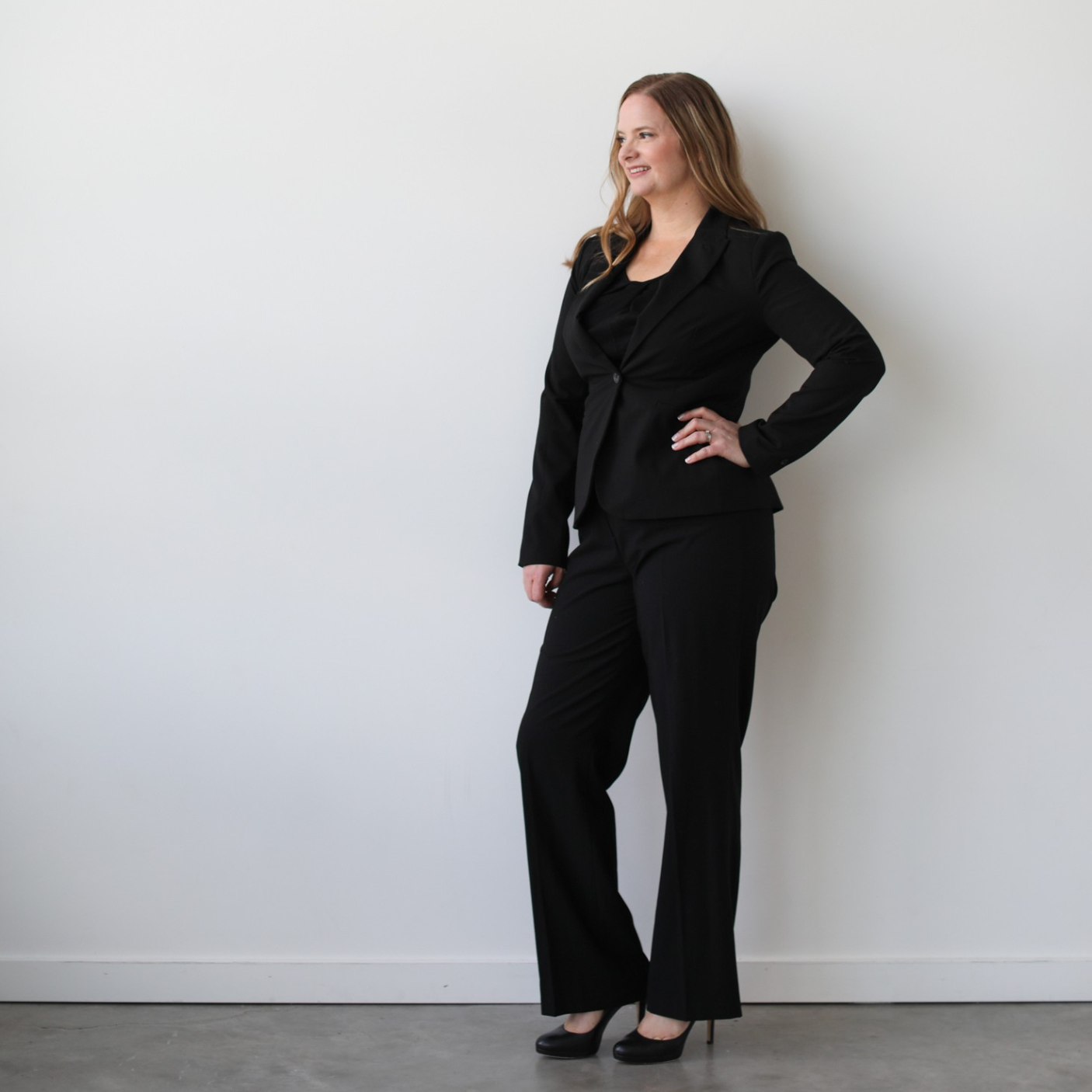 Jen Dunn Calgary Business Advisor in a black business suit standing against a plain white wall, with her hand on her hip, smiling slightly, wearing high heels.