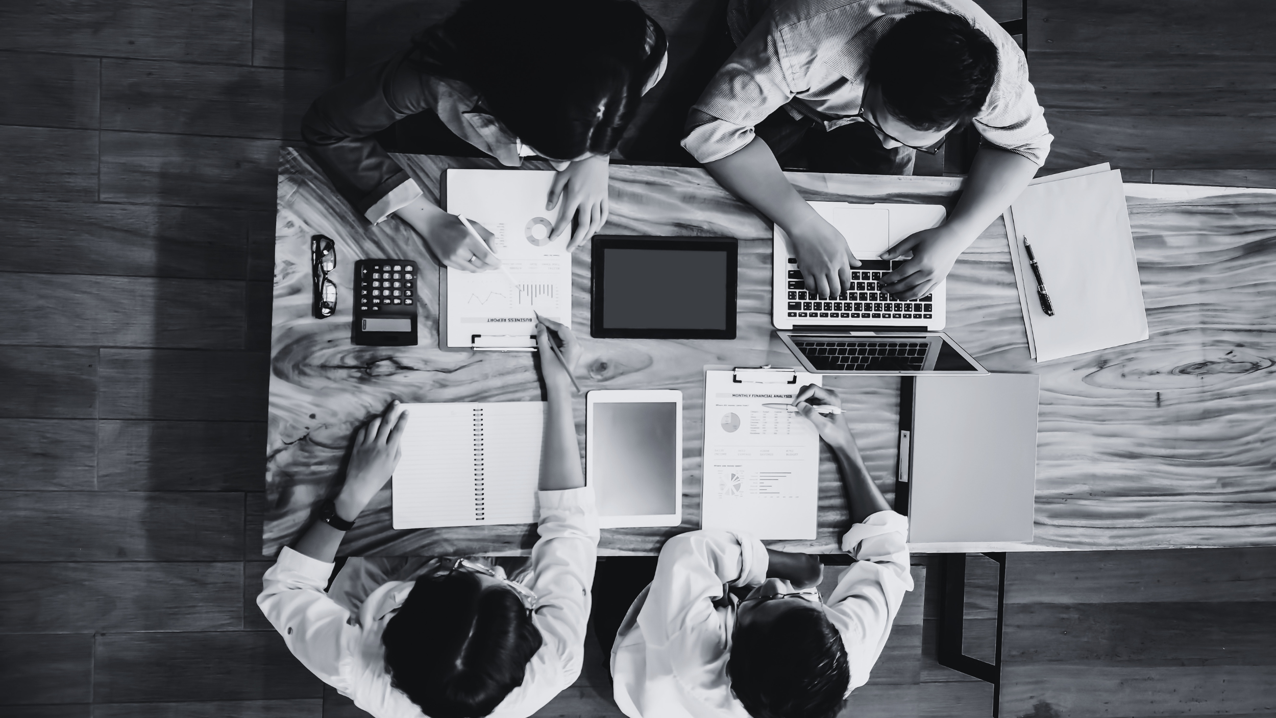 Top-down view of six people working at a wooden table with laptops, tablets, notebooks, pens, documents, glasses, and calculators, engaged in a business or teamwork meeting.
