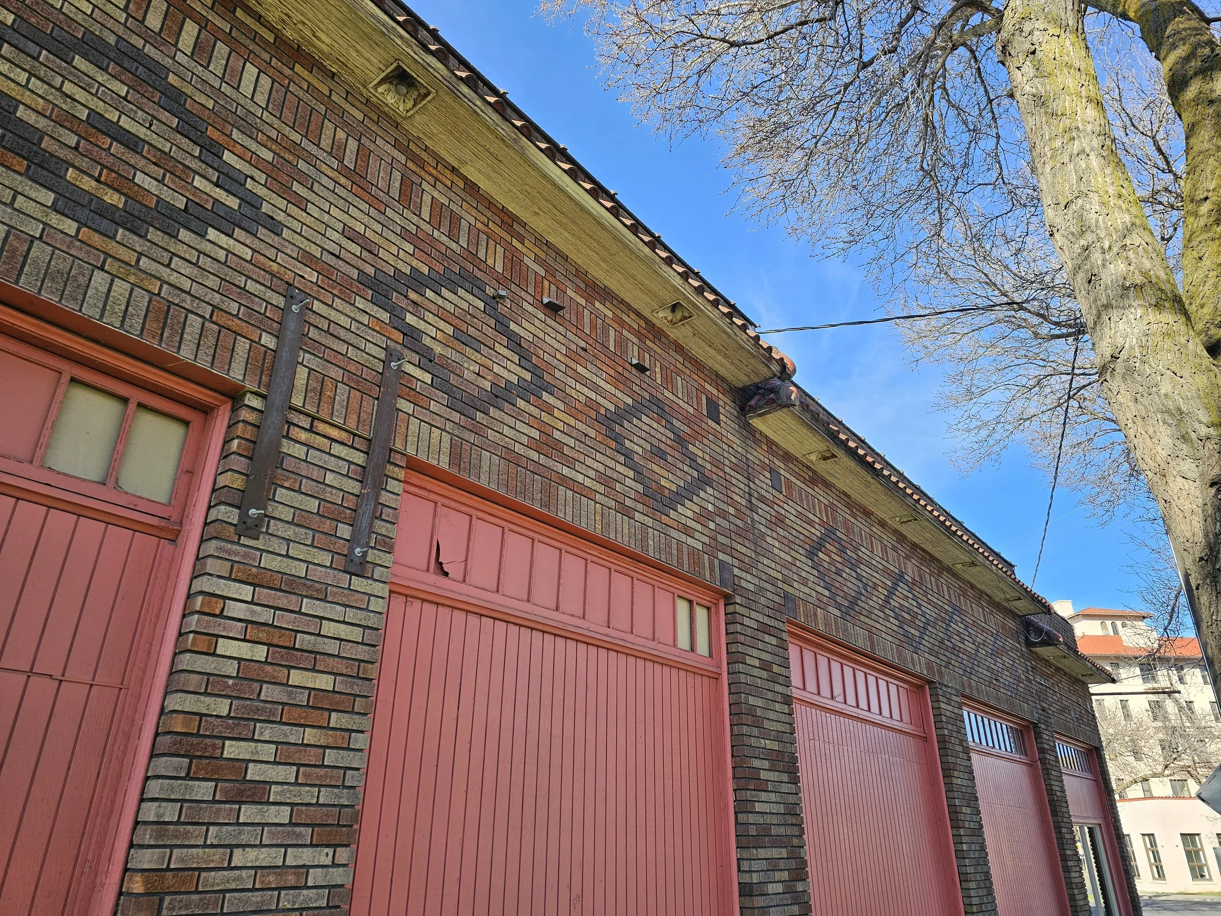 Brick building with red garage doors and a large tree with bare branches in front, under a blue sky.