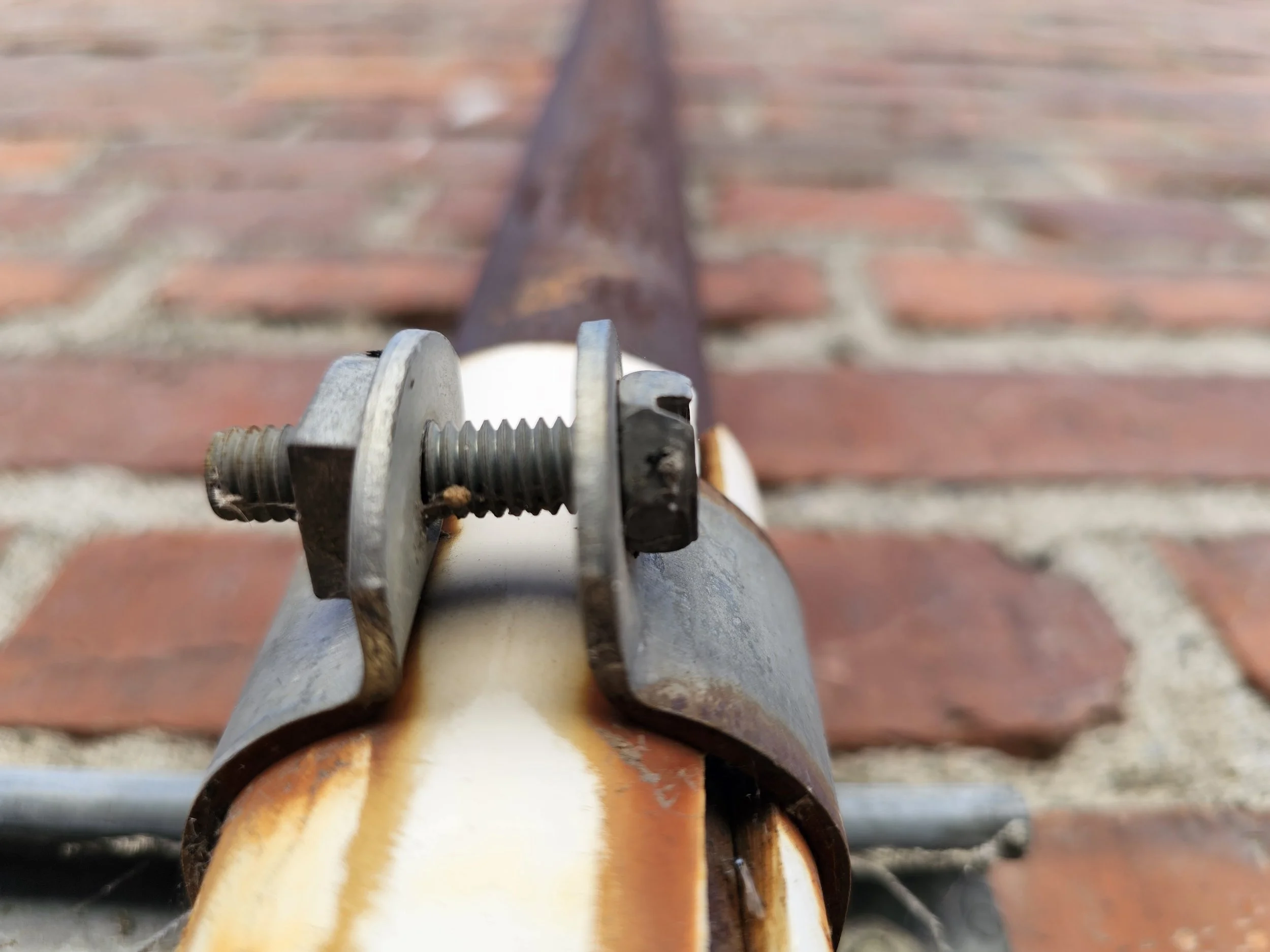 Close-up of a metal bolt and clamp on a rusty pole, with a brick-paved surface in the background.