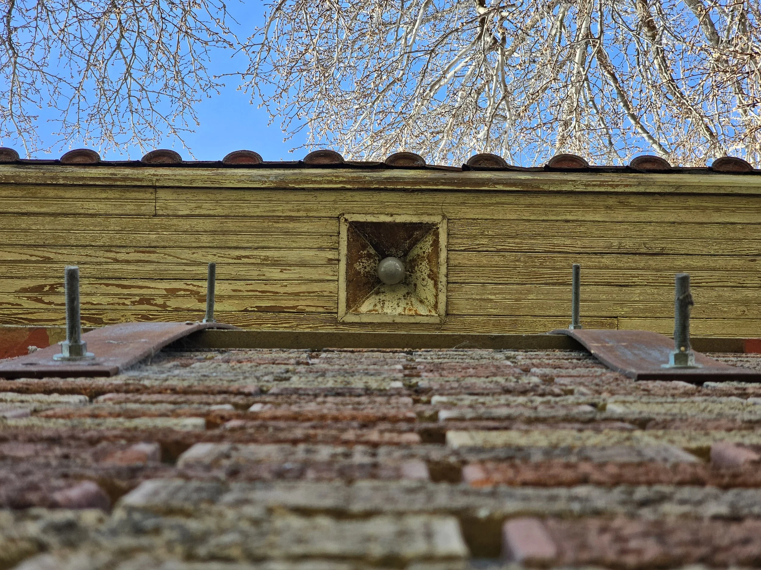 Upward view of the corner of a brick building with a metal bracket and a wooden section above it, a roof with red tiles, and trees with bare branches against a blue sky.