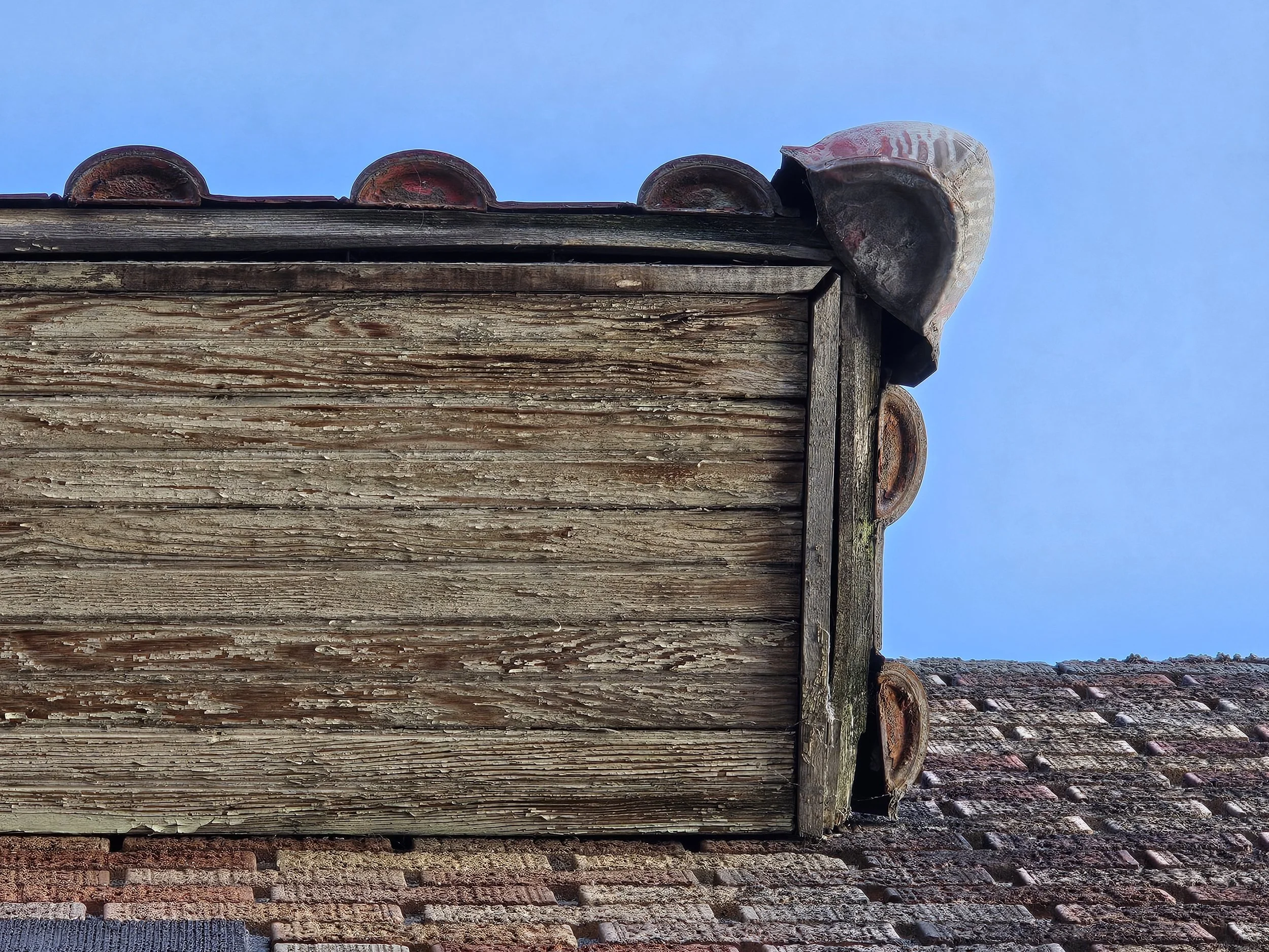 Close-up of an old, weathered wooden roof eave with clay tiles, showing peeling paint and moss, against a clear blue sky and part of a brick wall.