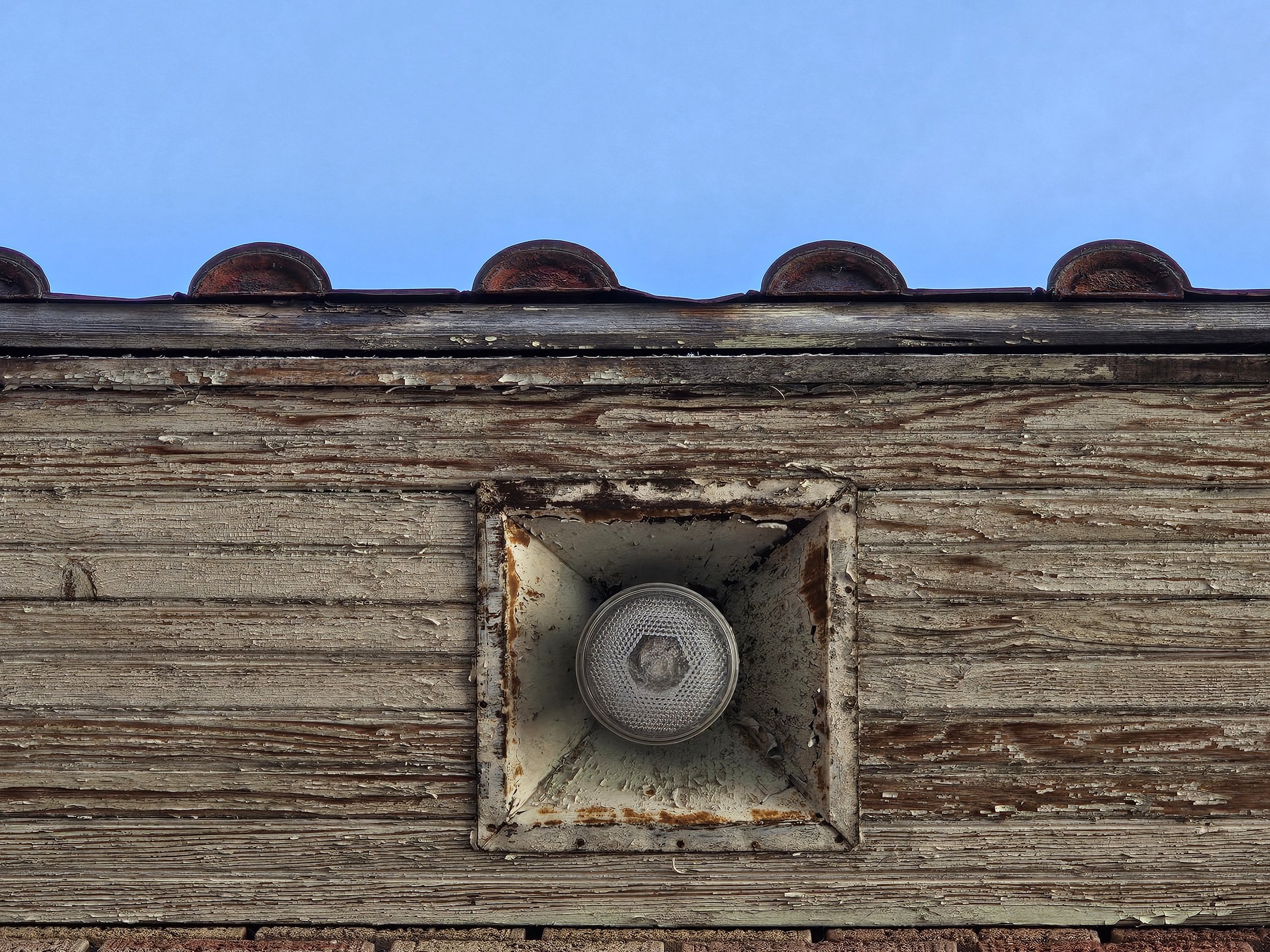 Close-up of a weathered wooden wall with a round outdoor light fixture in a rusted, square metal frame, and part of a tiled roof against a blue sky.