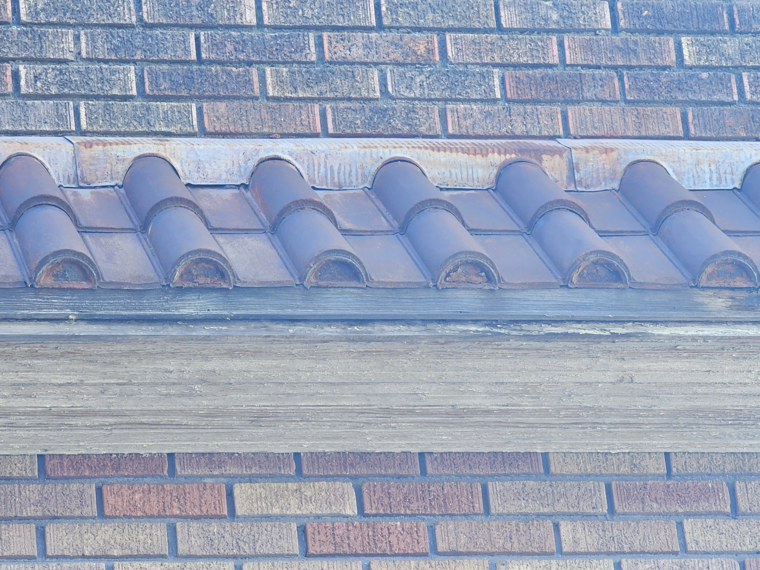 Close-up of a roof with curved tiles, a metal flashing strip above the tiles, and brick walls above and below.