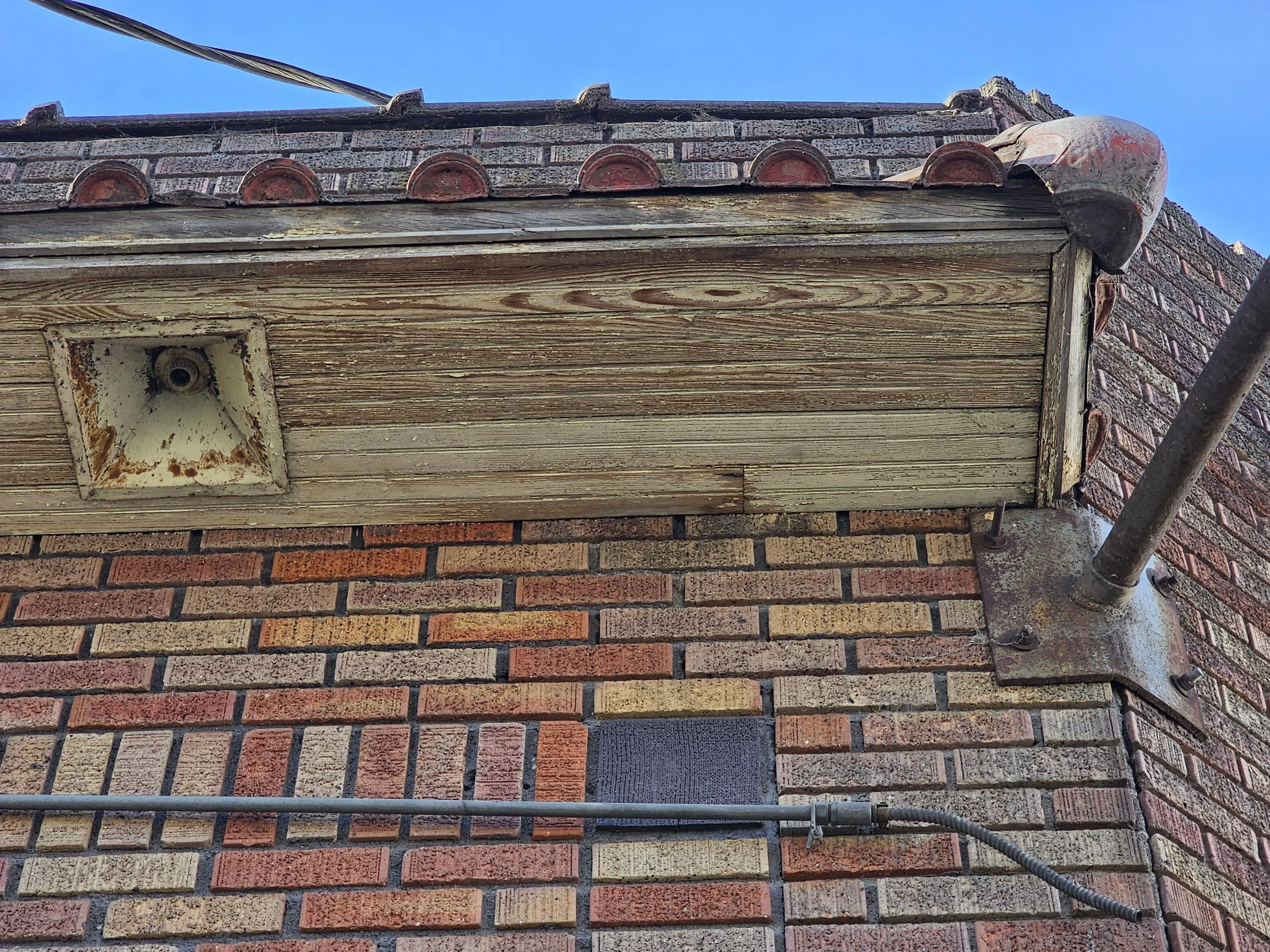 Close-up of a brick building corner with weathered wood soffit, an old light fixture, and metal support brackets.