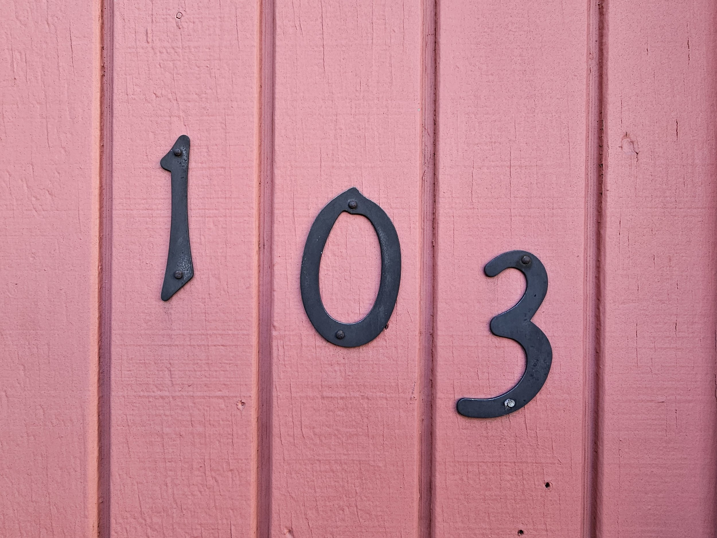 Number 103 on a pink wooden wall, with black metal numbers.