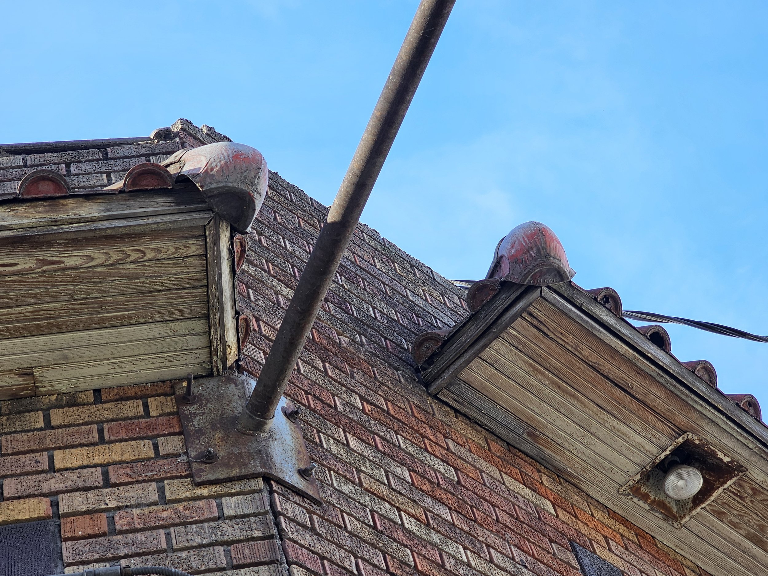 Close-up of an old brick building corner with a rusted metal pipe and a wooden overhang with a light fixture, under a blue sky.