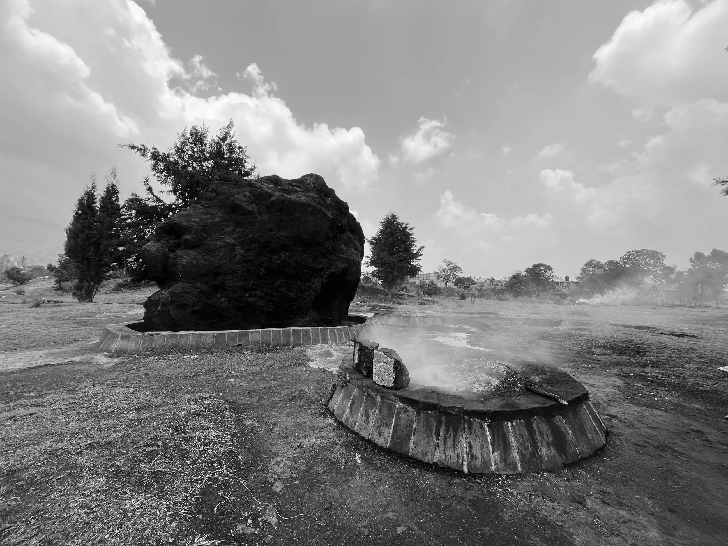 A large boulder on a circular concrete base in an open landscape with trees and buildings in the background, and steam rising from a hot spring or geothermal feature in the foreground, all in black and white.