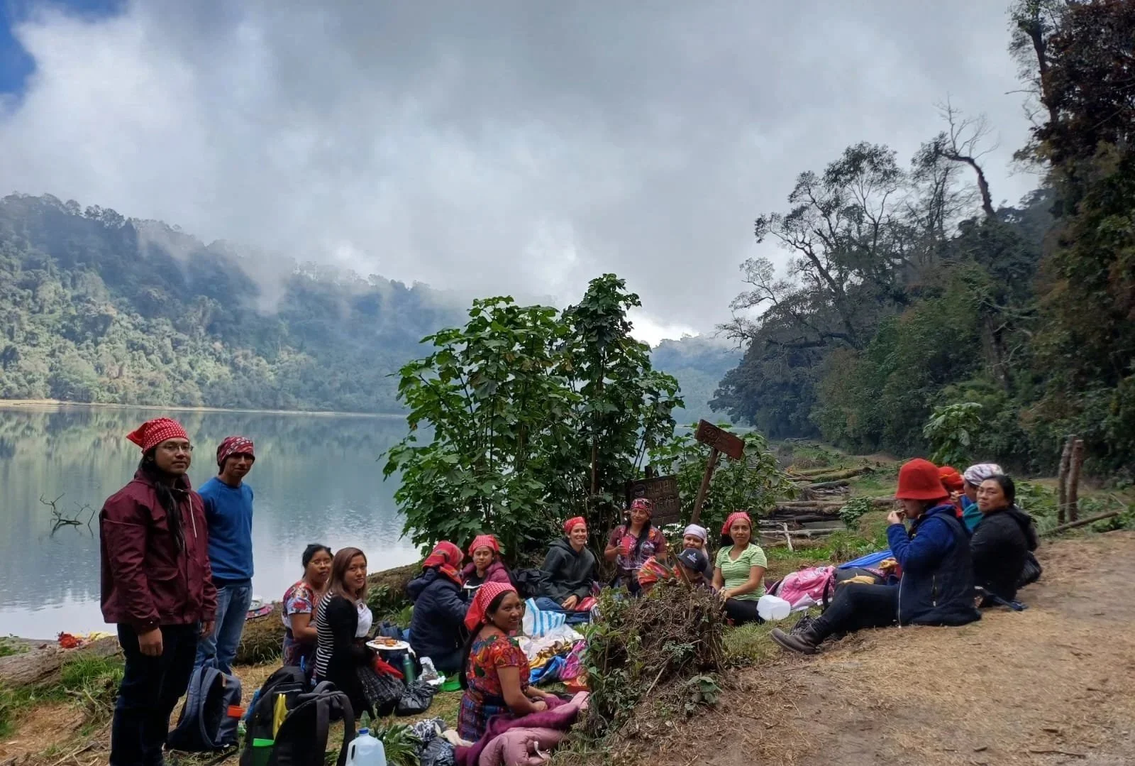 Group of people sitting and standing along a lakeshore, surrounded by trees and mountains, with cloudy sky overhead.