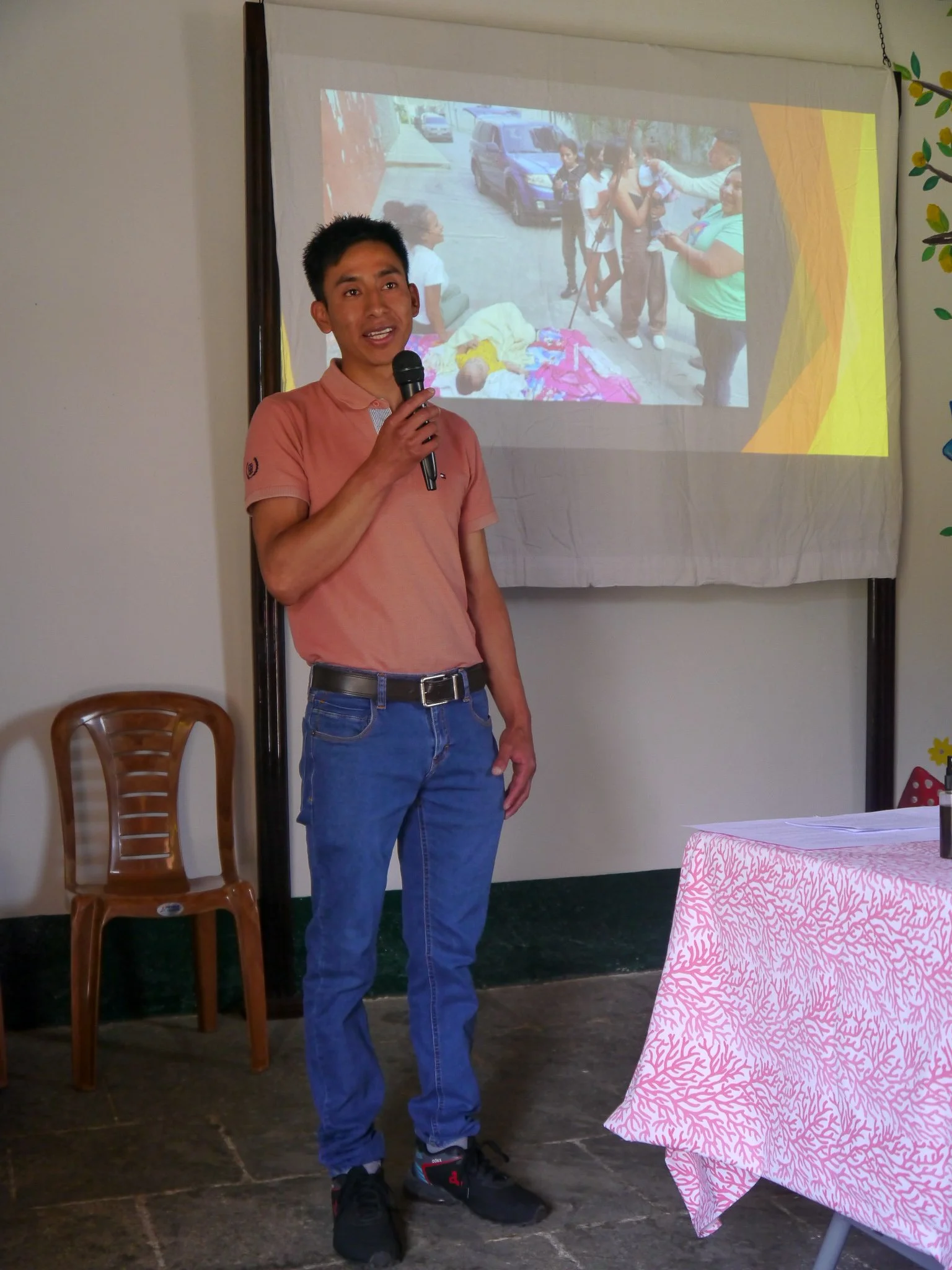 A young man in a pink polo shirt and jeans is speaking into a microphone, standing in front of a projection screen showing a photo of people outdoors, with parked cars and a group of individuals, some lying on the ground, and others standing and talk