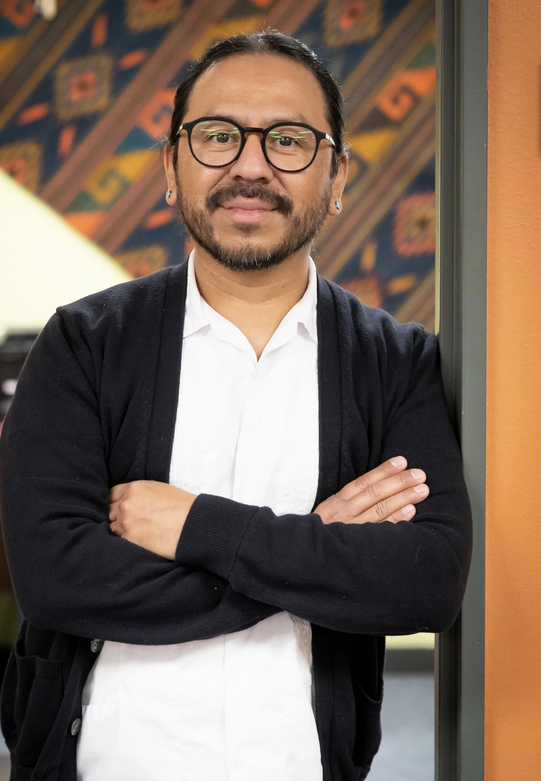 A man with glasses, a beard and long hair tied back, wearing a white shirt and black cardigan, standing with arms crossed next to an orange wall, in front of a colorful geometric background.