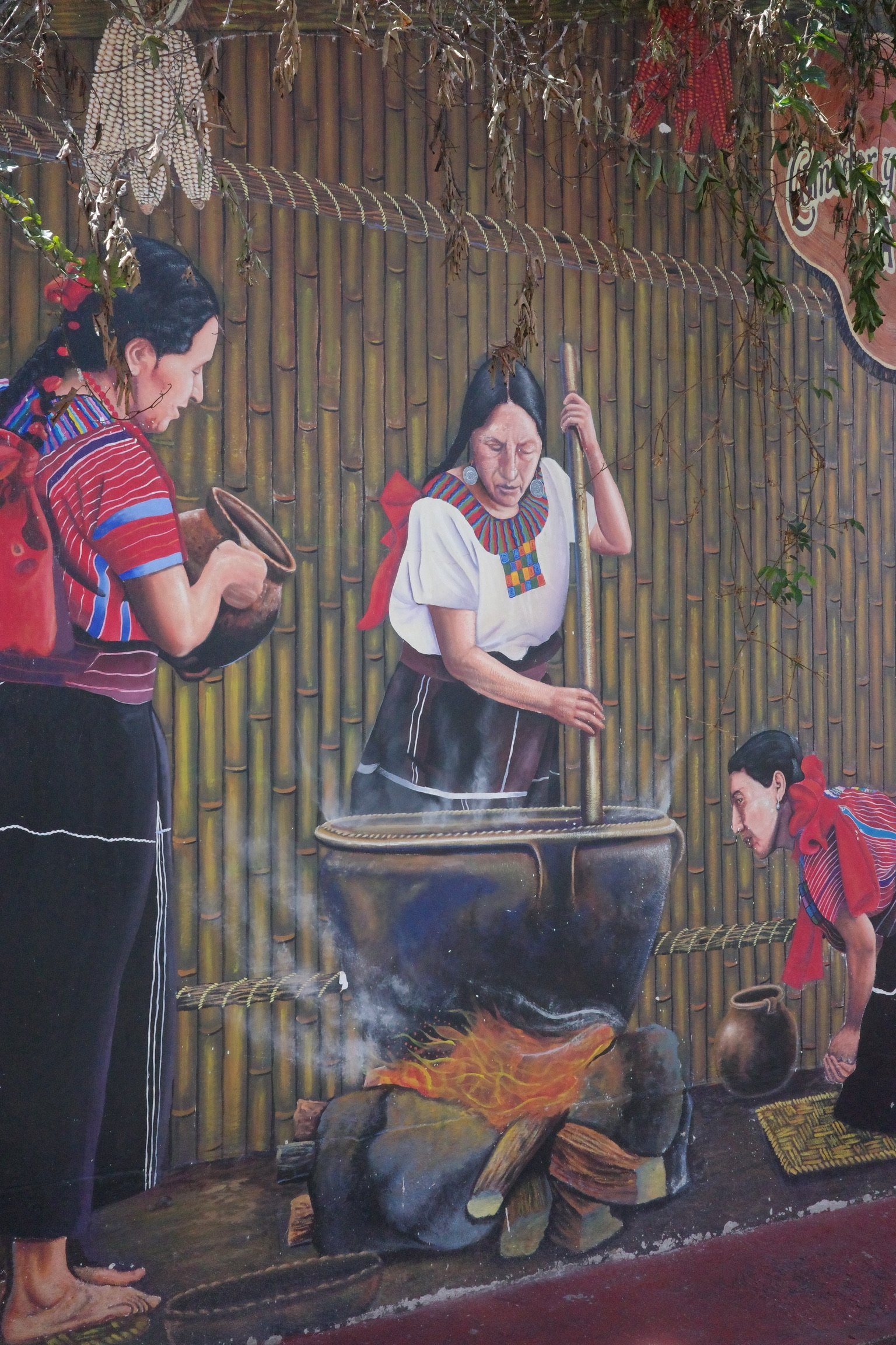 Mural depicting women in traditional clothing preparing something over a fire with a large pot, bamboo wall background.