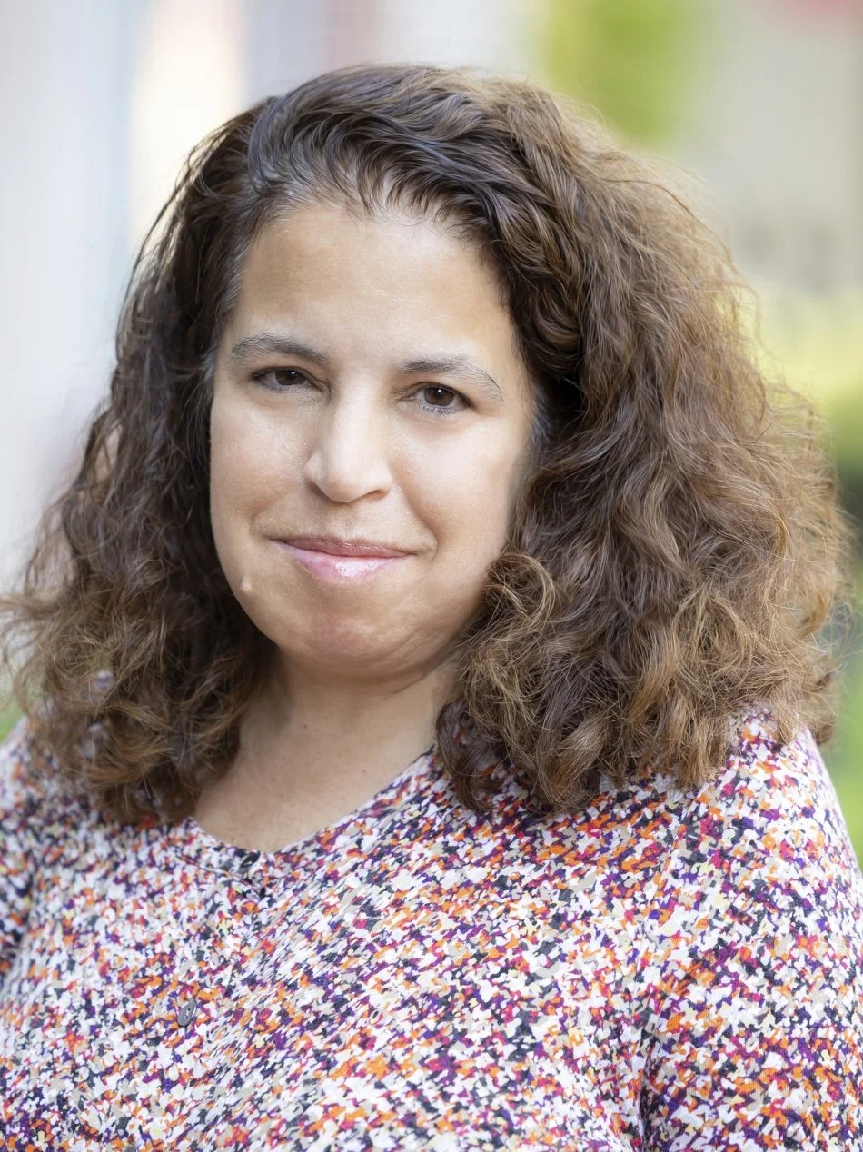 Close-up of a woman with curly brown hair, wearing a multicolored patterned top, smiling slightly, outdoors with blurred background.