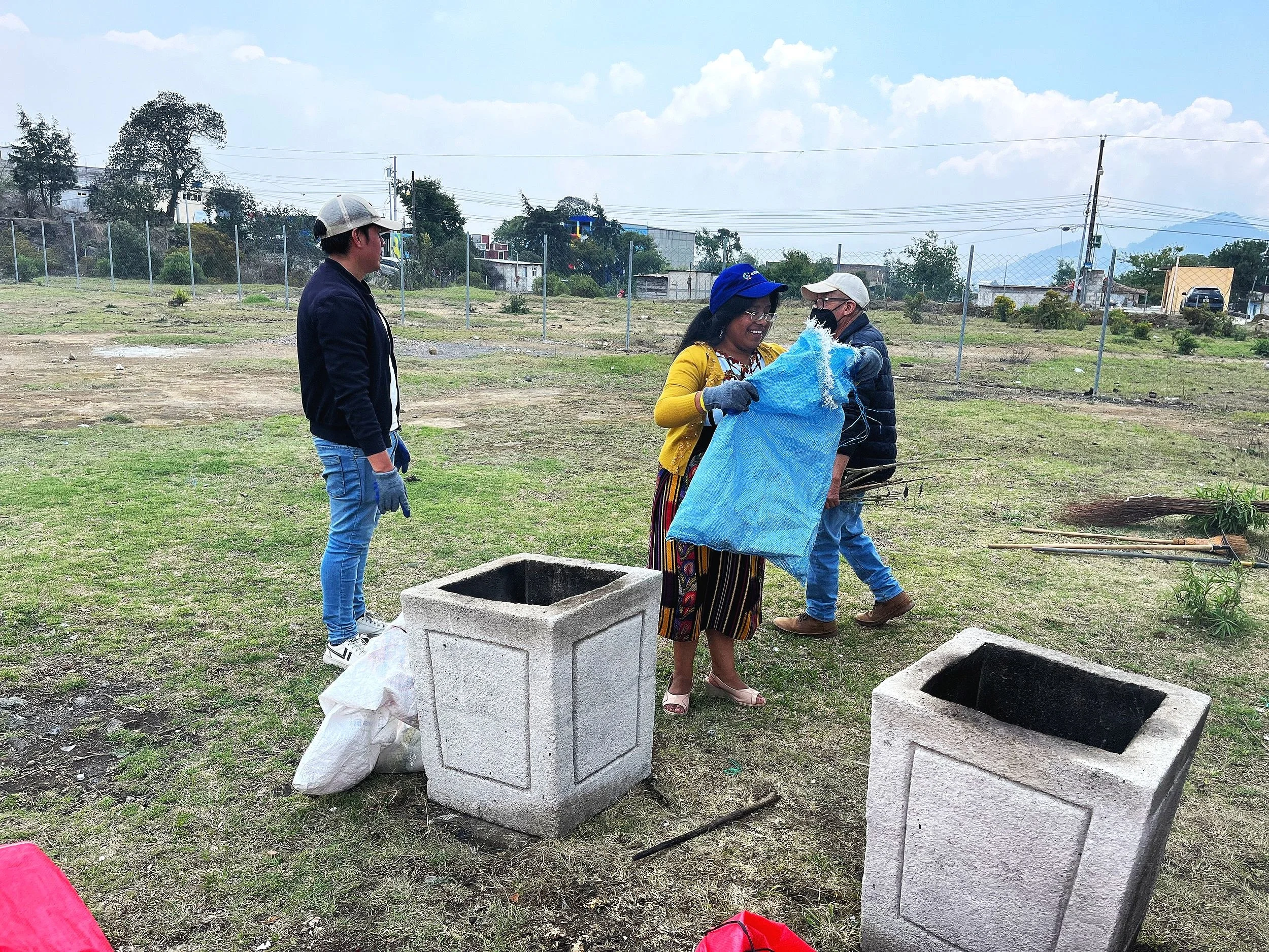 Four people working on a community cleanup project outdoors, two women and two men, with empty concrete trash receptacles in the foreground and open grassy field in the background.