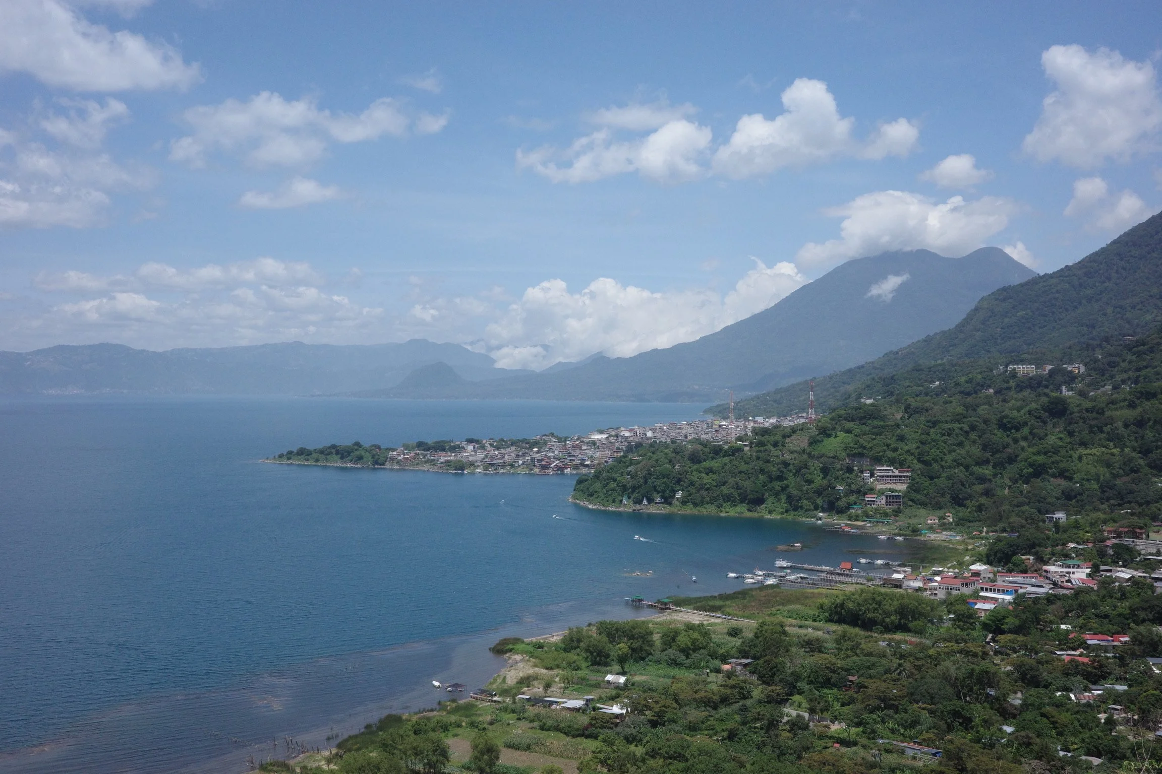 Scenic view of a lake surrounded by green hills and mountains, with a small town along the shoreline, under a partly cloudy sky.