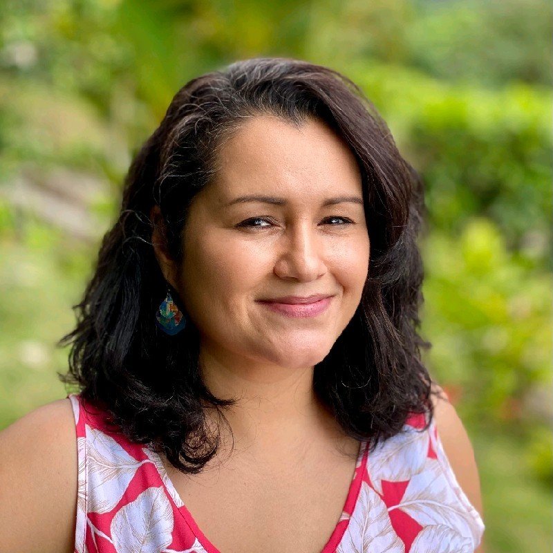 A woman with dark curly hair smiling outdoors, wearing a sleeveless top with a white and pink floral pattern, and earrings.