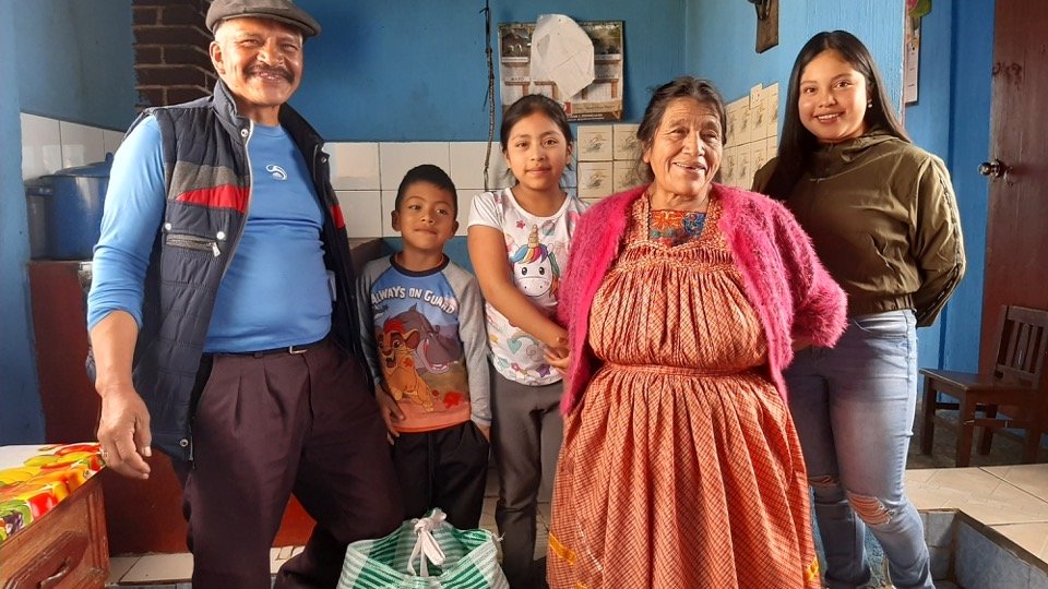 A group of five people, including two men and three young girls, standing inside a room with blue walls and tiled parts. The older woman in the center is wearing traditional clothing and a pink shawl. The others are casually dressed, and everyone is smiling.