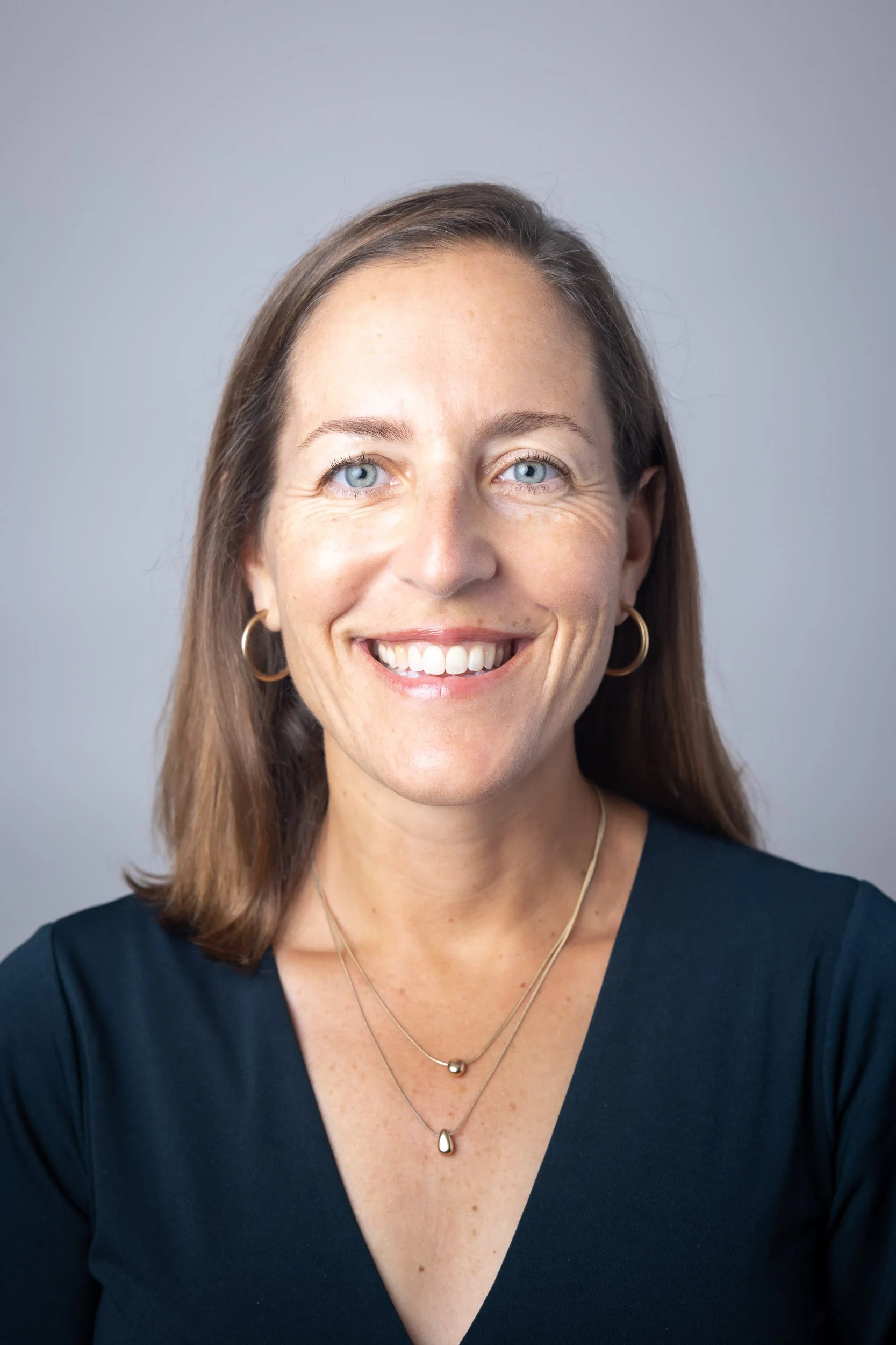 A smiling woman with shoulder-length brown hair, blue eyes, wearing hoop earrings and layered necklaces, dressed in a dark V-neck top, against a plain gray background.