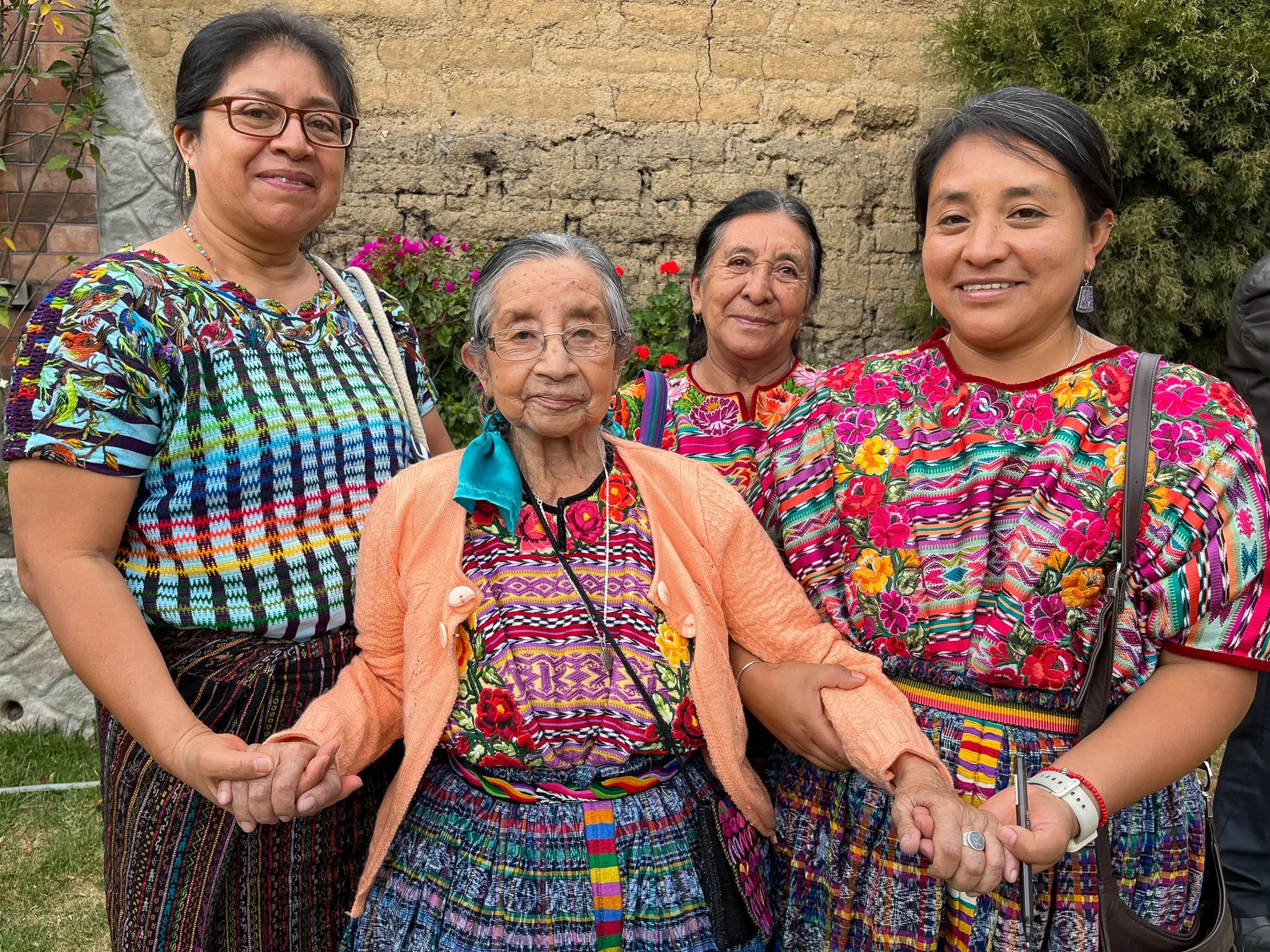 Four women in colorful traditional Mexican clothing standing outdoors, holding hands, with a stone and brick wall and trees in the background.