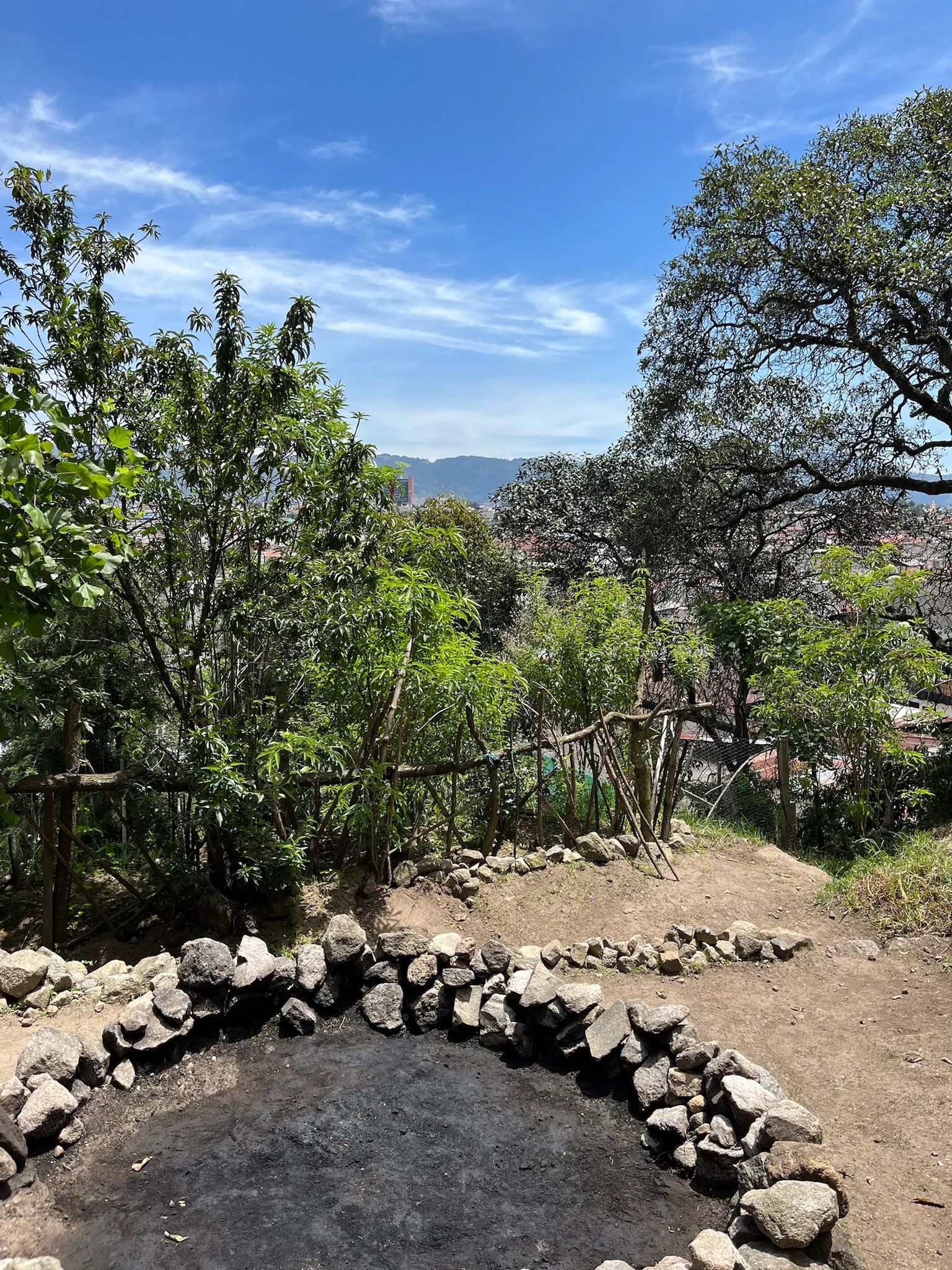 View of a garden with a circular stone fire pit, surrounded by trees, with a view of distant mountains and a partly cloudy blue sky.