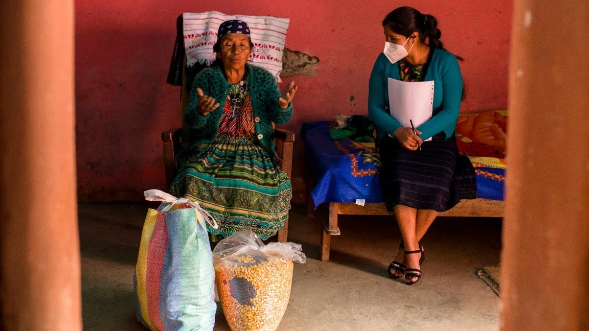 An elderly woman and a younger woman wearing a face mask sitting in a room with pink walls. The elderly woman is seated on a wooden chair with a pillow behind her head, gesturing with her hands. The younger woman is sitting on a small bed, holding a 