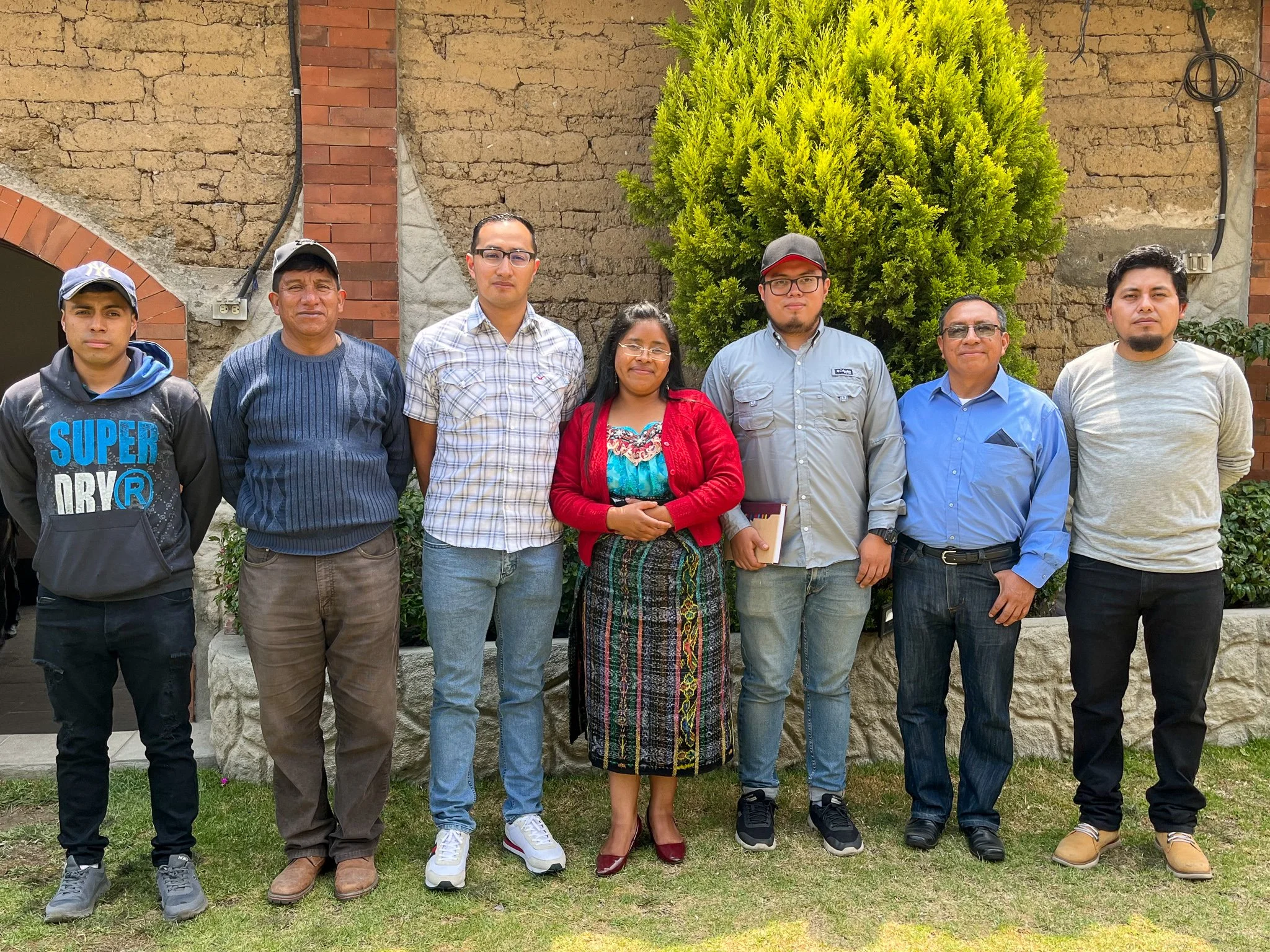 Group of eight people standing outdoors in front of a stone and brick wall with a tree, posing for a photo.