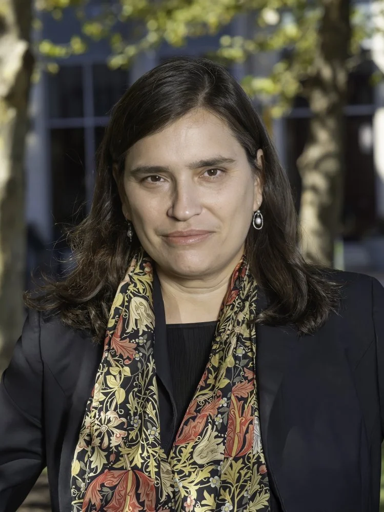 A woman with dark shoulder-length hair standing outdoors with trees and a building in the background.