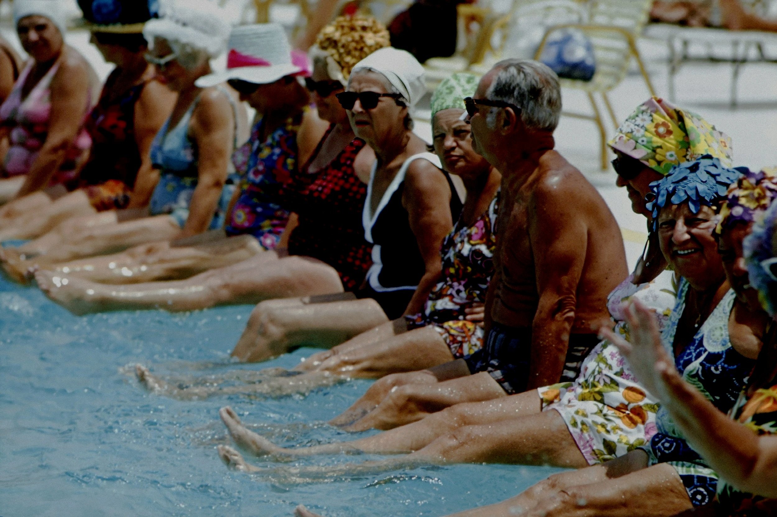 A group of elderly people sitting by the edge of a swimming pool with their feet in the water, wearing summer clothes and hats