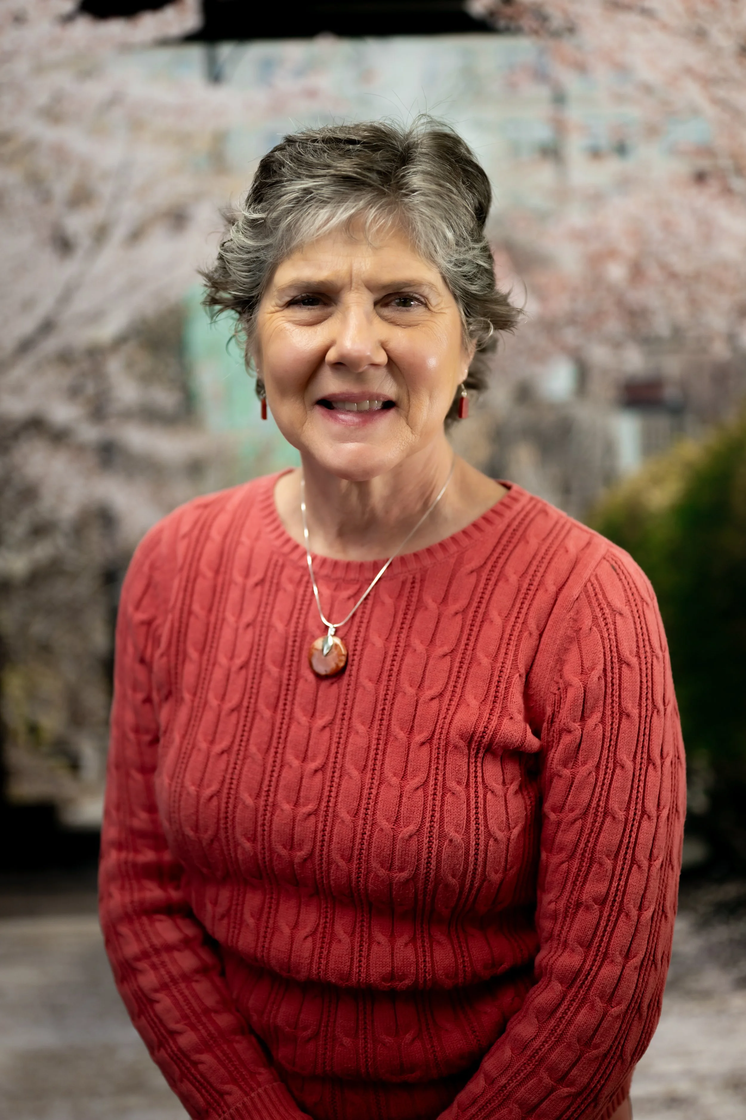 An older woman with short gray hair, wearing a red cable-knit sweater and a necklace with a stone pendant, smiling at the camera against a blurred background of cherry blossom trees and possibly a building.
