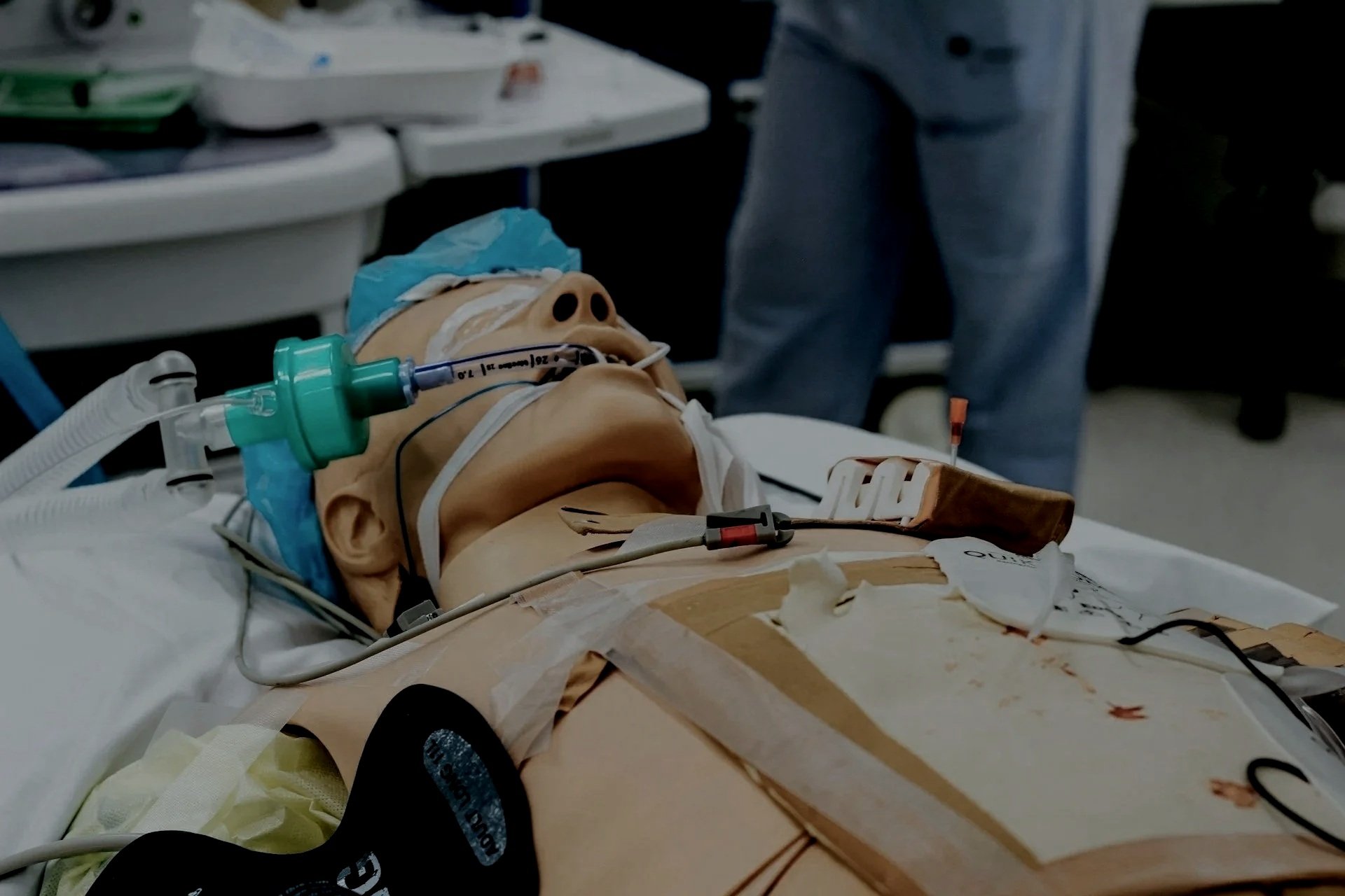 A medical simulation mannequin lying on a hospital bed with medical tubes and sensors attached.