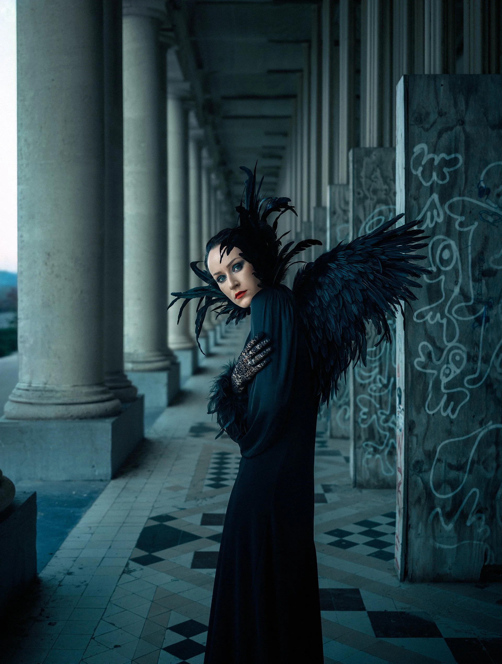 A woman dressed in a black costume with large feathered wings stands in a corridor with columns and graffiti-covered panels.
