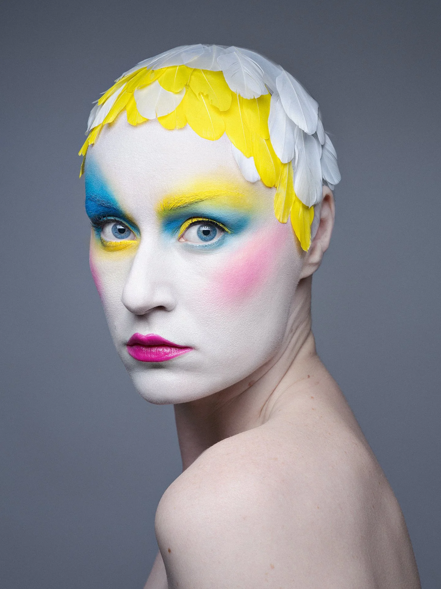 Close-up portrait of a woman with white skin, wearing vibrant yellow, blue, and pink makeup, and with white and yellow feathered hair accessories.