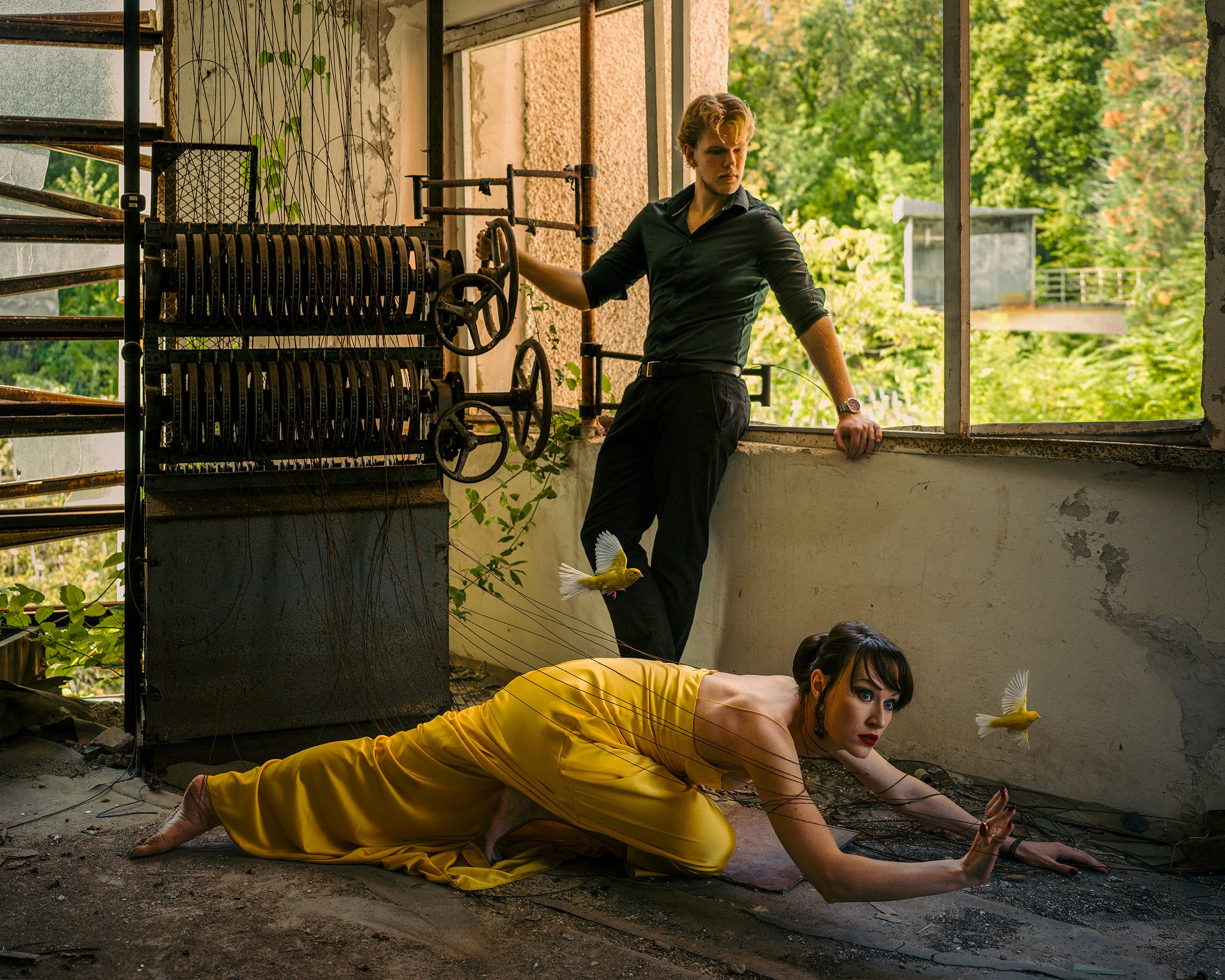 A woman in a yellow dress crawling on the ground with two birds flying nearby, and a man standing by an open window in an abandoned building.