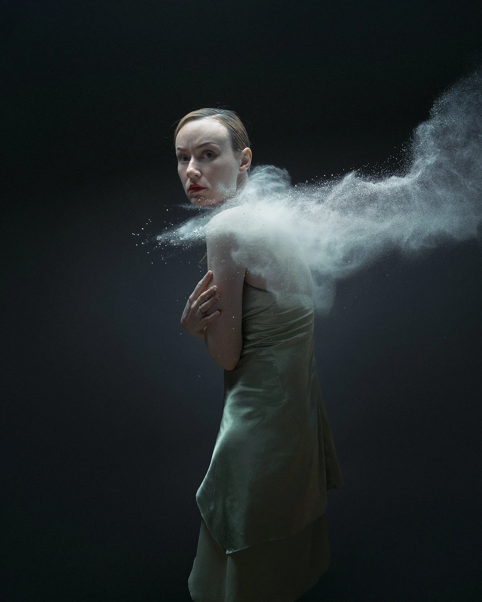 A woman with short hair and red lipstick standing in front of a black background, with white powder or dust swirling around her, creating a dynamic effect.