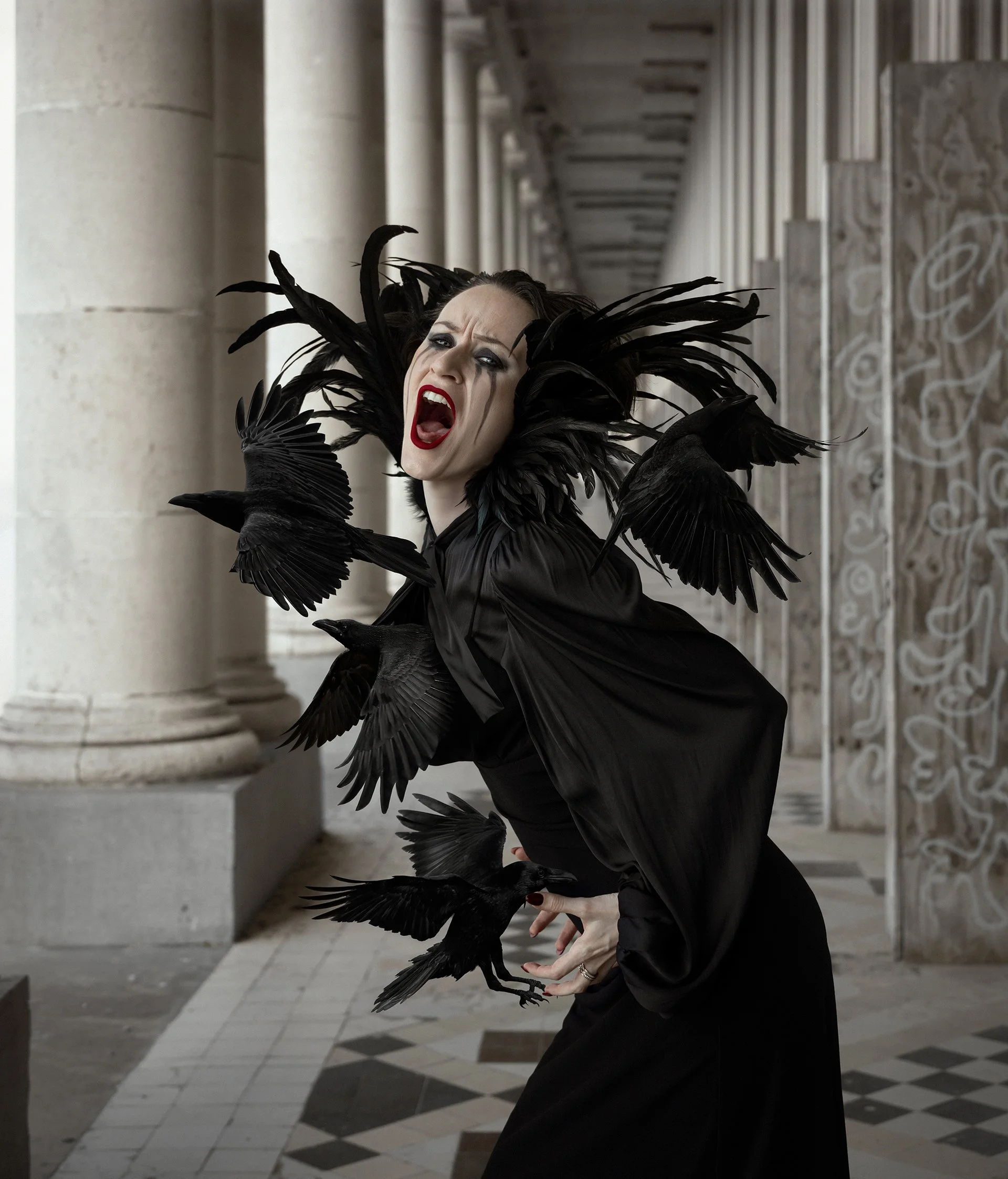 A woman dressed in black with dramatic makeup and black feathers on her head, surrounded by black birds, stands in a classical architectural setting.