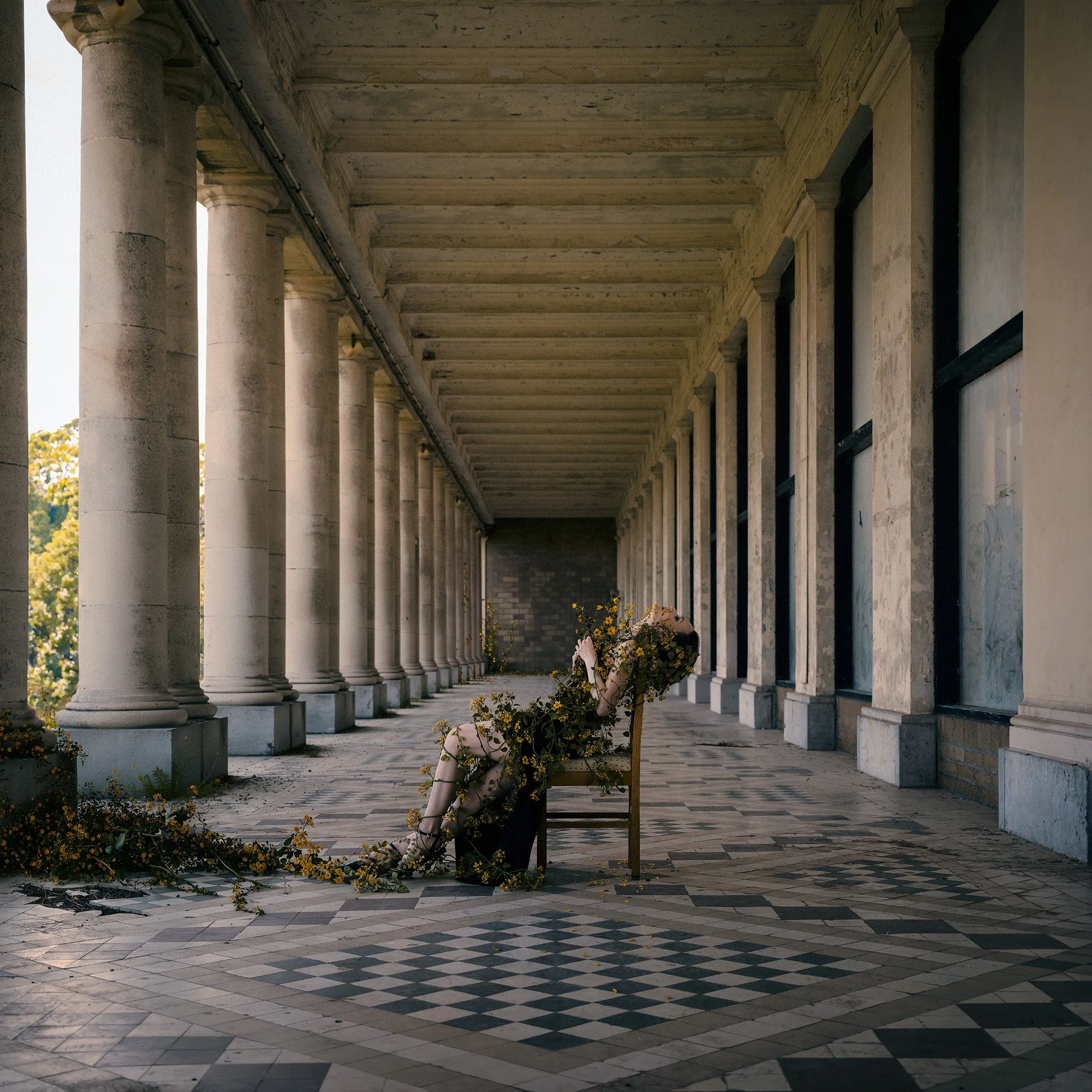 A person wearing a black dress is reclining on a wooden chair on a patterned tiled floor, covered in yellow flowers, on an outdoor marble-columned veranda.