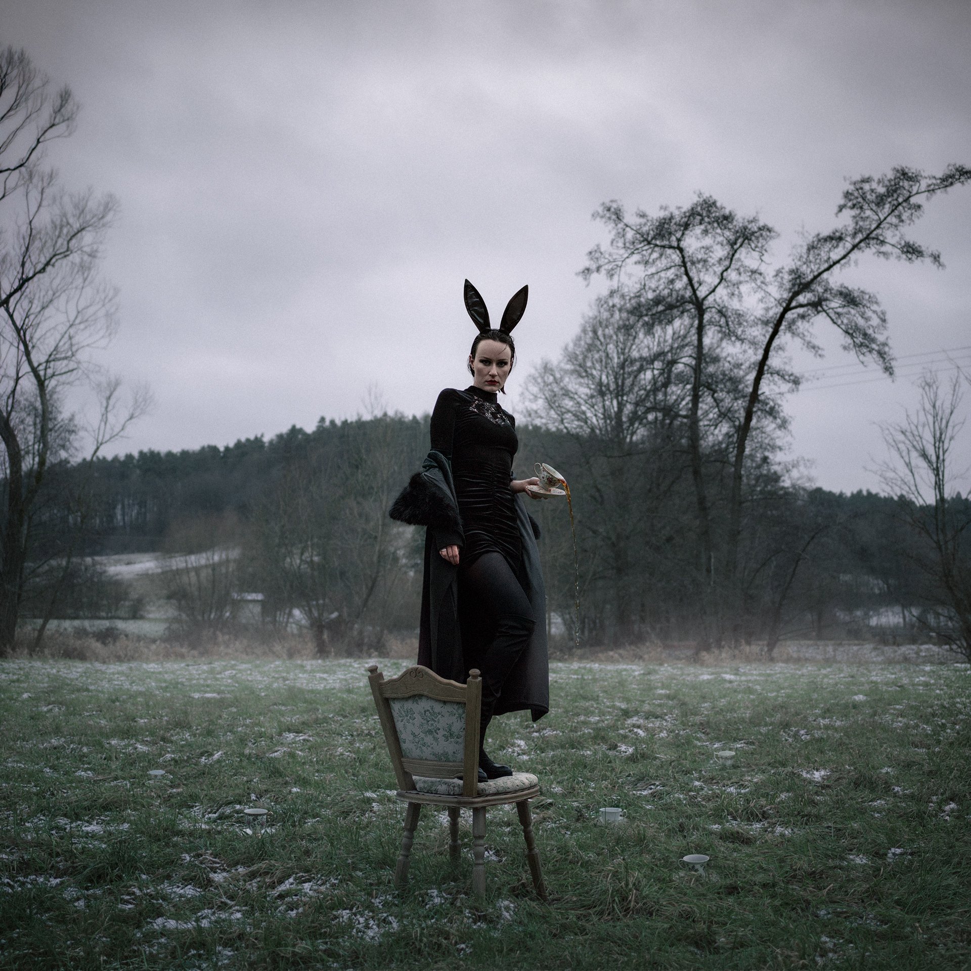 A woman dressed in black with bunny ears standing on a chair in an outdoor, overcast landscape holding a pipe.