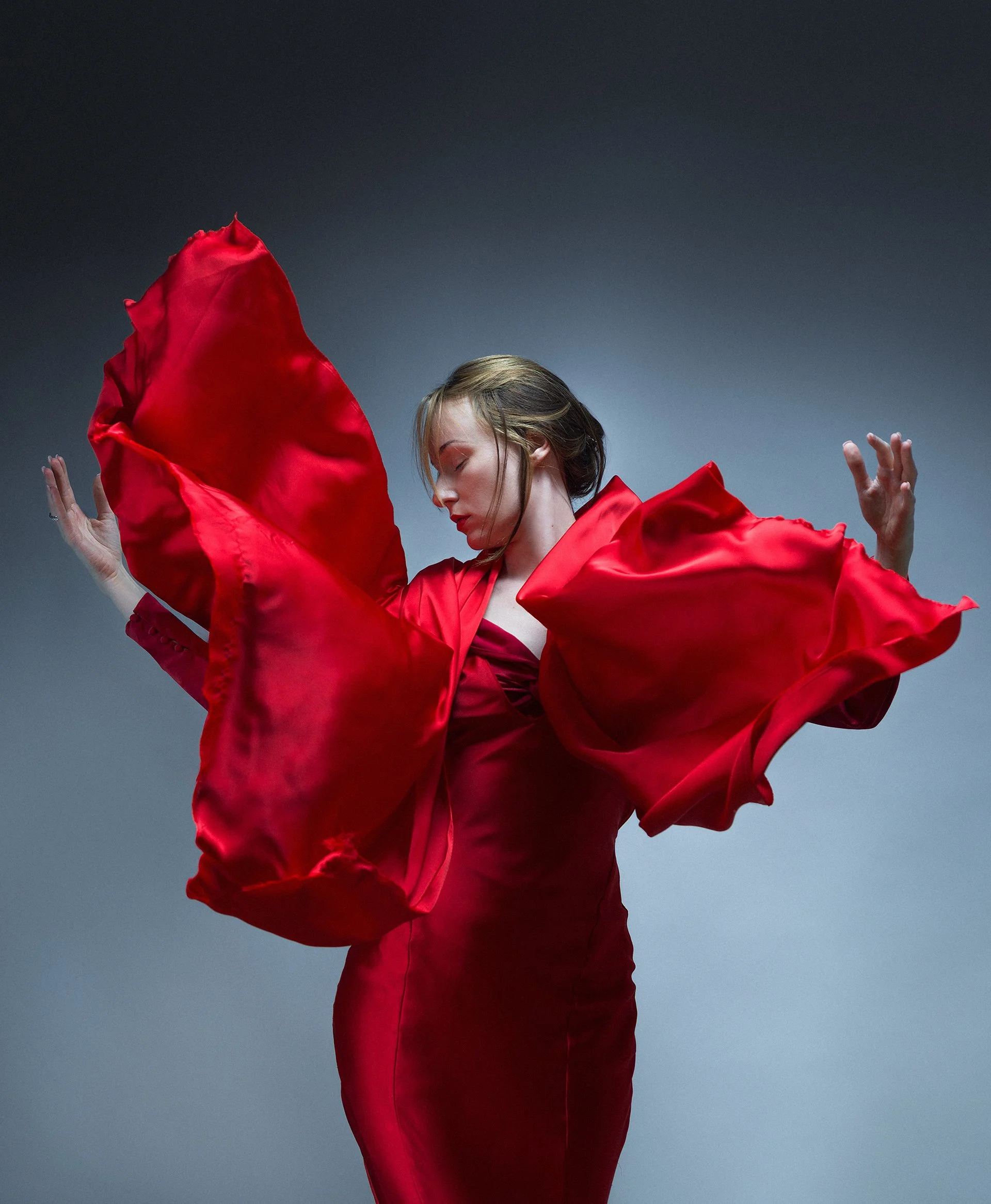 A woman with short blonde hair wearing a flowing red dress, with her eyes closed and arms raised, appears to be dancing or performing against a gradient gray background.