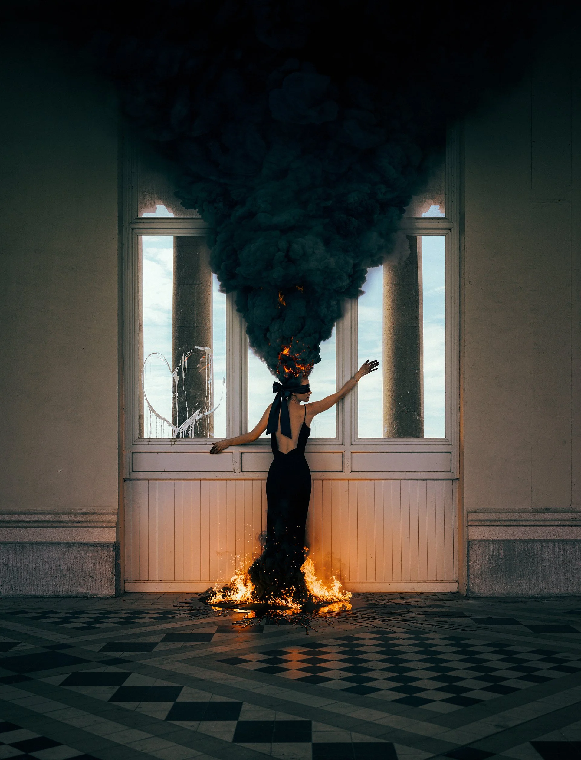 A woman in a black dress with a blindfold is standing in a room with her arms outstretched. Her head is on fire and emitting thick black smoke that rises upward. The woman is also standing in a circle of fire on the floor in front of her, with a shat