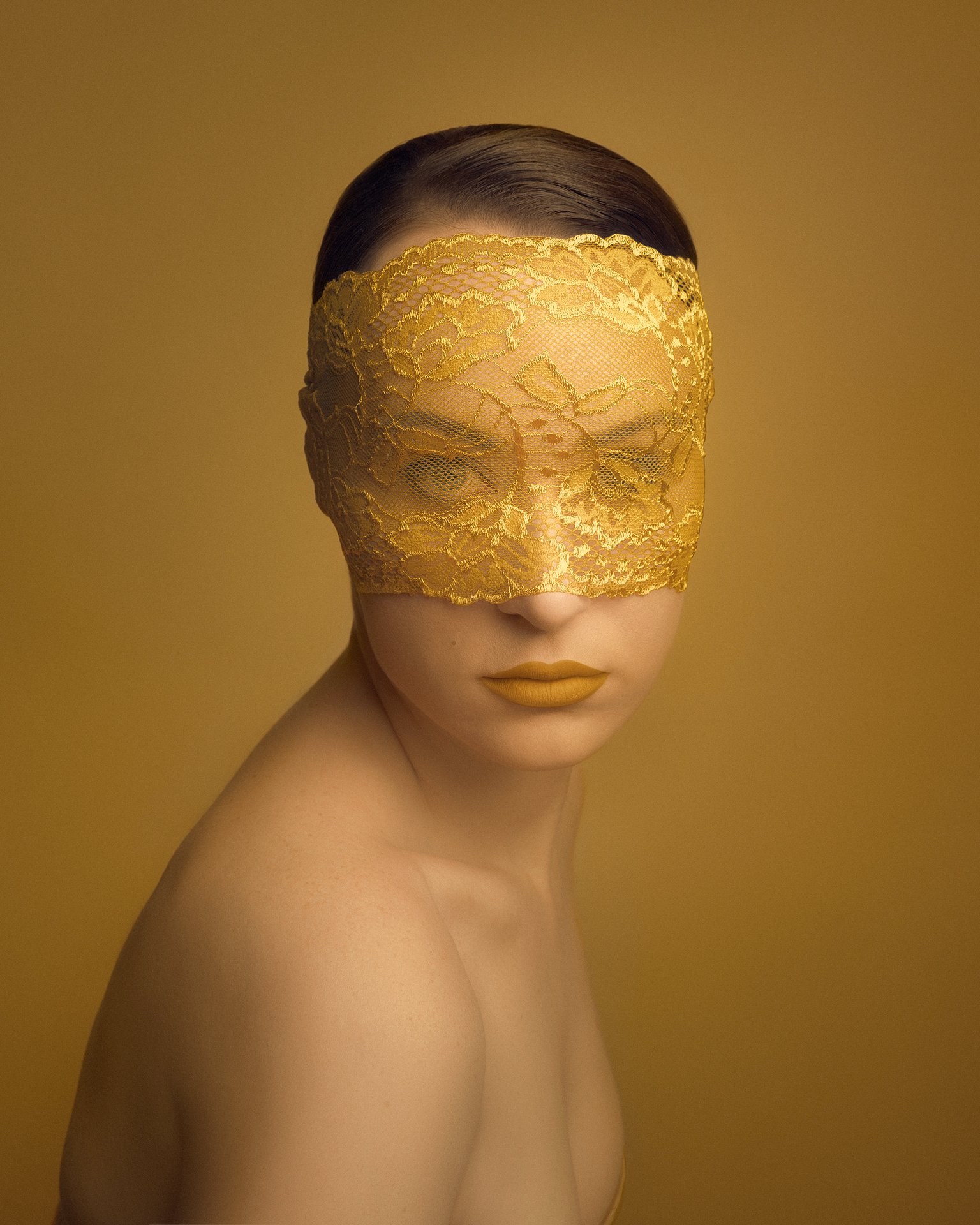 A woman with smooth skin and dark hair pulled back, wearing yellow lipstick, poses against a yellow background. She has a delicate yellow lace mask covering her eyes and face, creating a mysterious and elegant look.