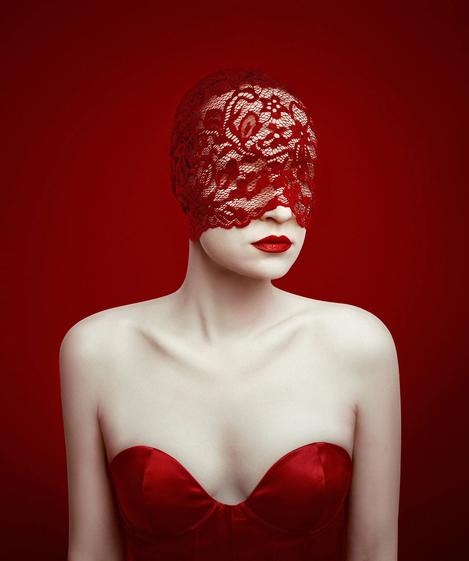A woman with pale skin and red lipstick, wearing a red strapless dress and a red lace mask covering her eyes and forehead, against a red background.